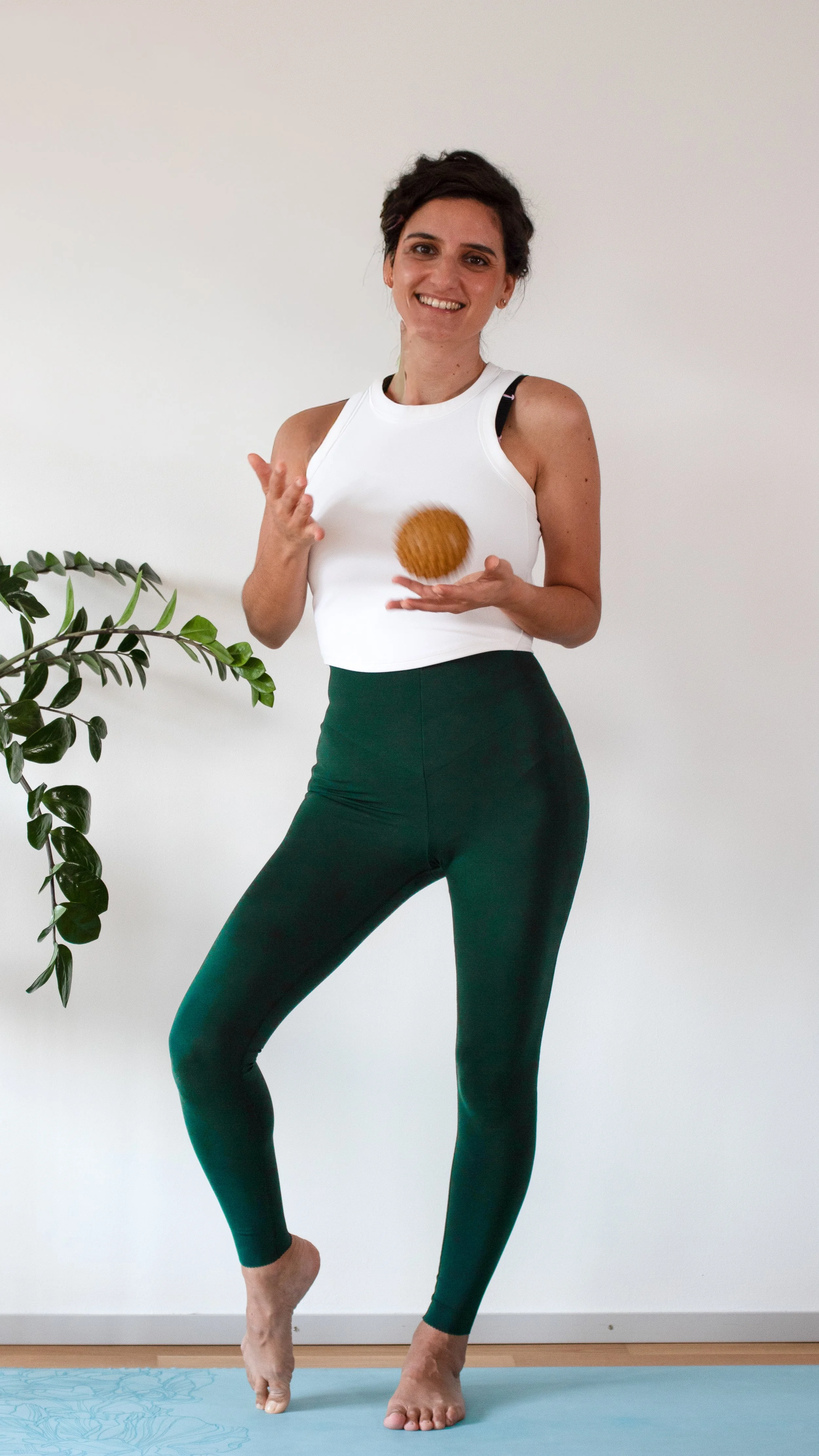 Woman in workout attire standing on a yoga mat, smiling, holding a wooden massage ball in her right hand, with a plant visible on the left side.