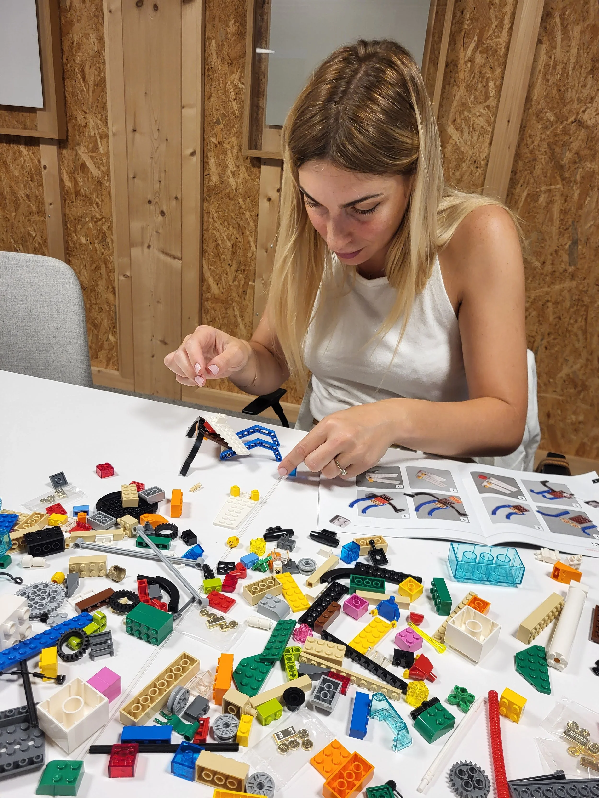 A woman assembling a LEGO set at a white table with various LEGO pieces and an instruction manual.
