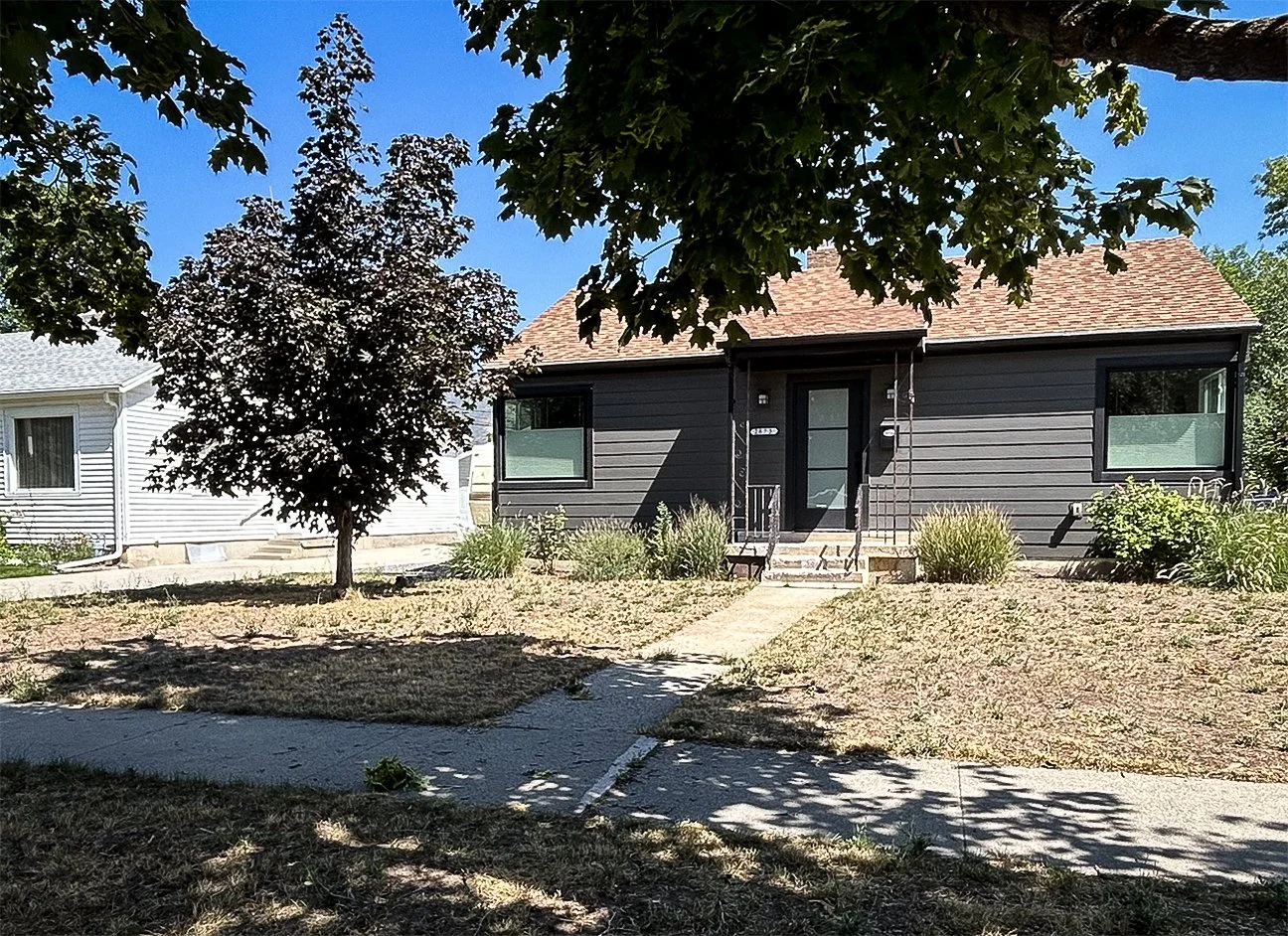 Front view of a black house with a red-tiled roof, with a sidewalk leading up to the front door, and a tree in the yard, under a clear blue sky.