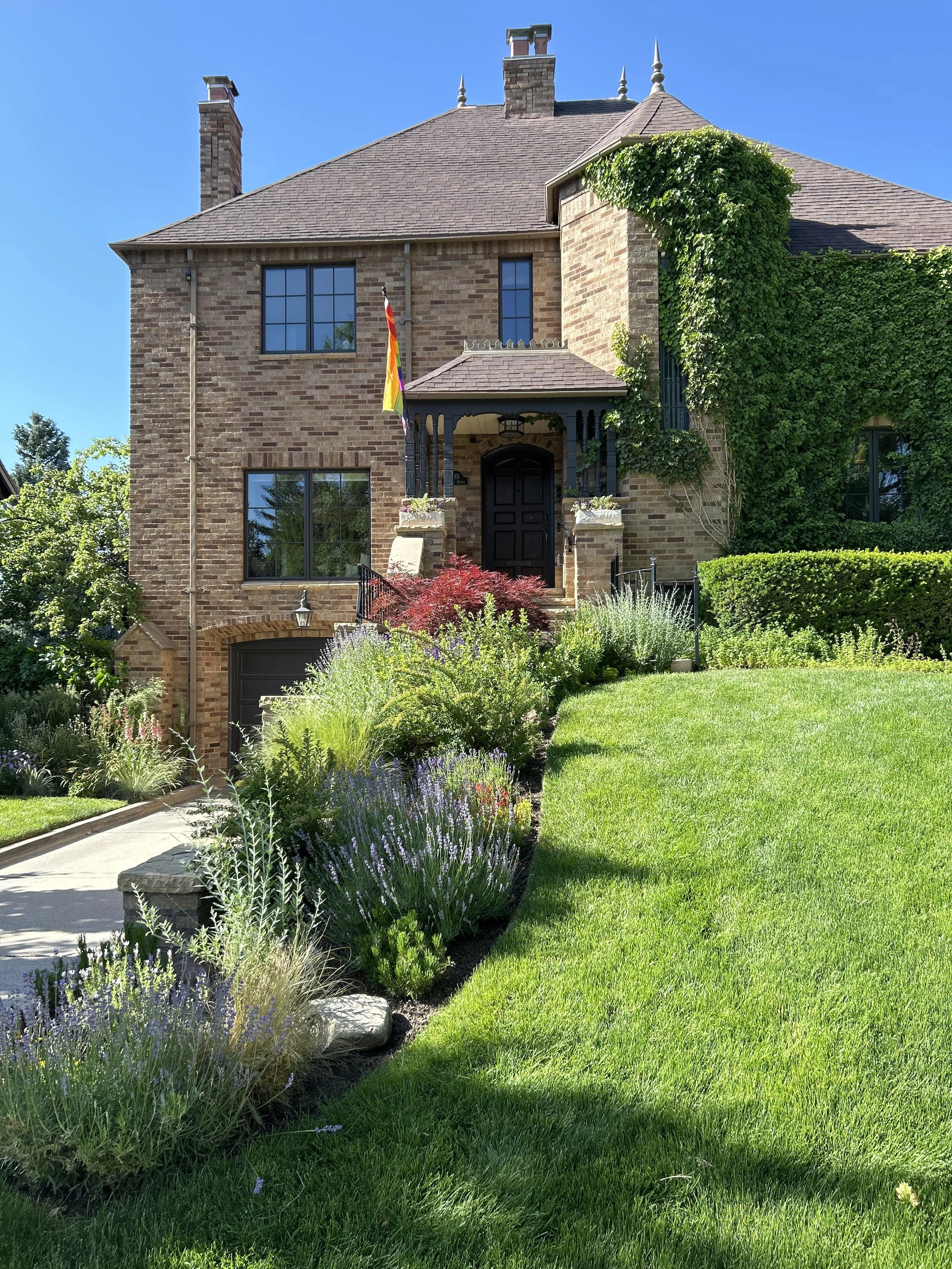 A two-story brick house with an ivy-covered side, a dark front door, a rainbow flag, and lush green landscaped garden with bushes and flowering plants in front.
