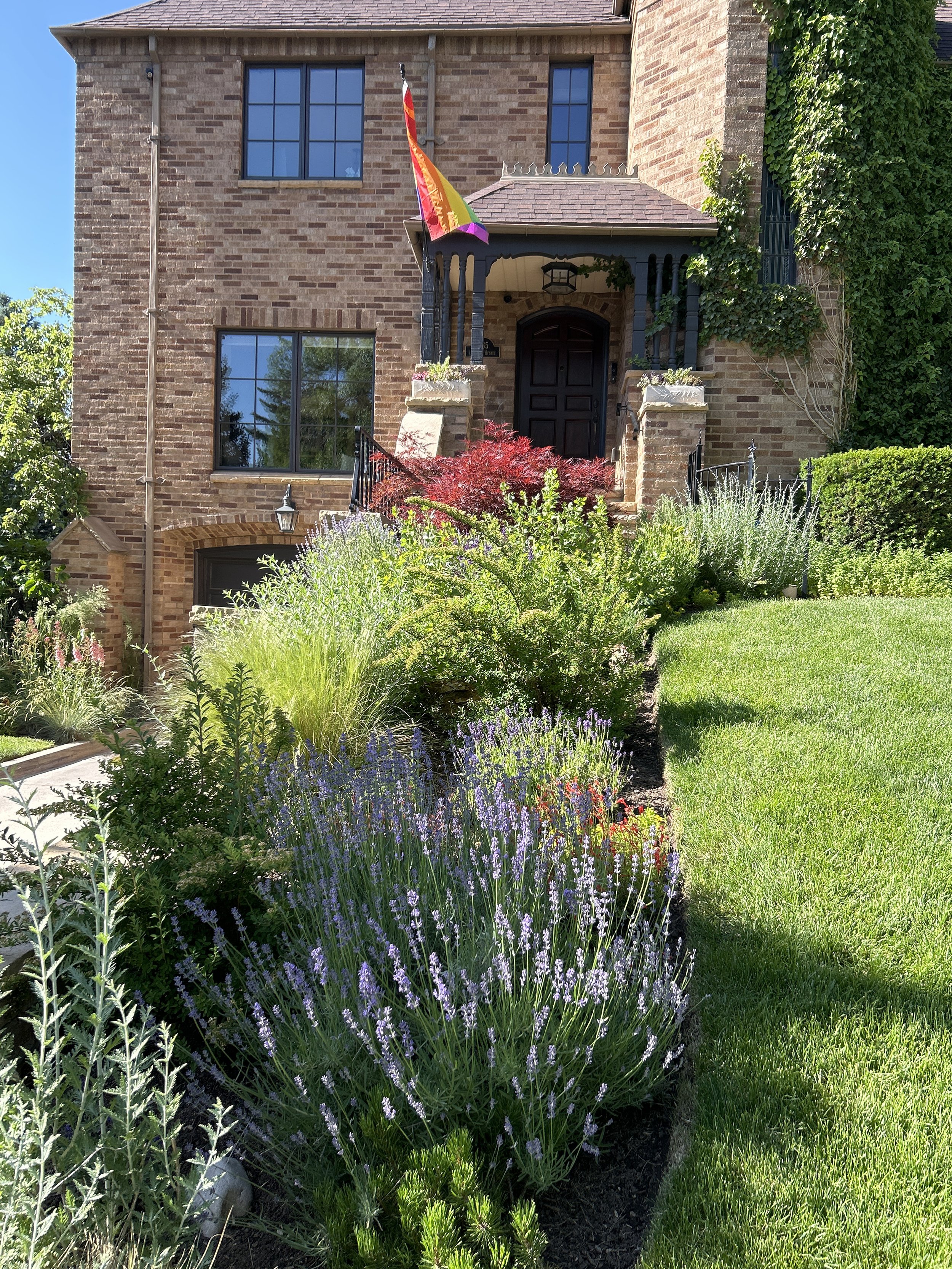 Front view of a brick house with a landscaped front yard. There is a rainbow pride flag hanging near the door, surrounded by various green plants and purple flowers.