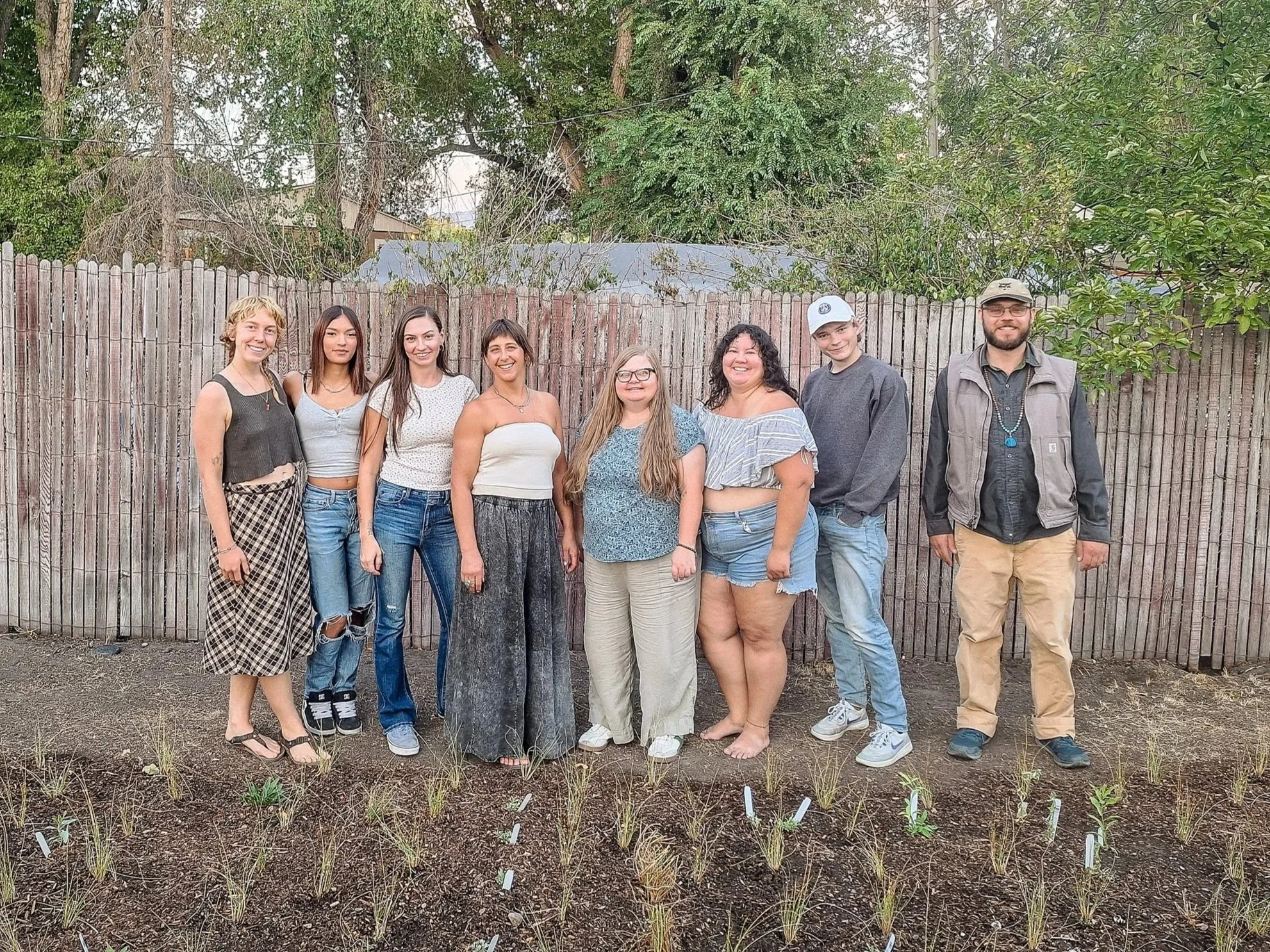 A group of nine people standing outdoors in front of a wooden fence with trees in the background. They are smiling and standing next to a garden bed with small plants marked with white tags.