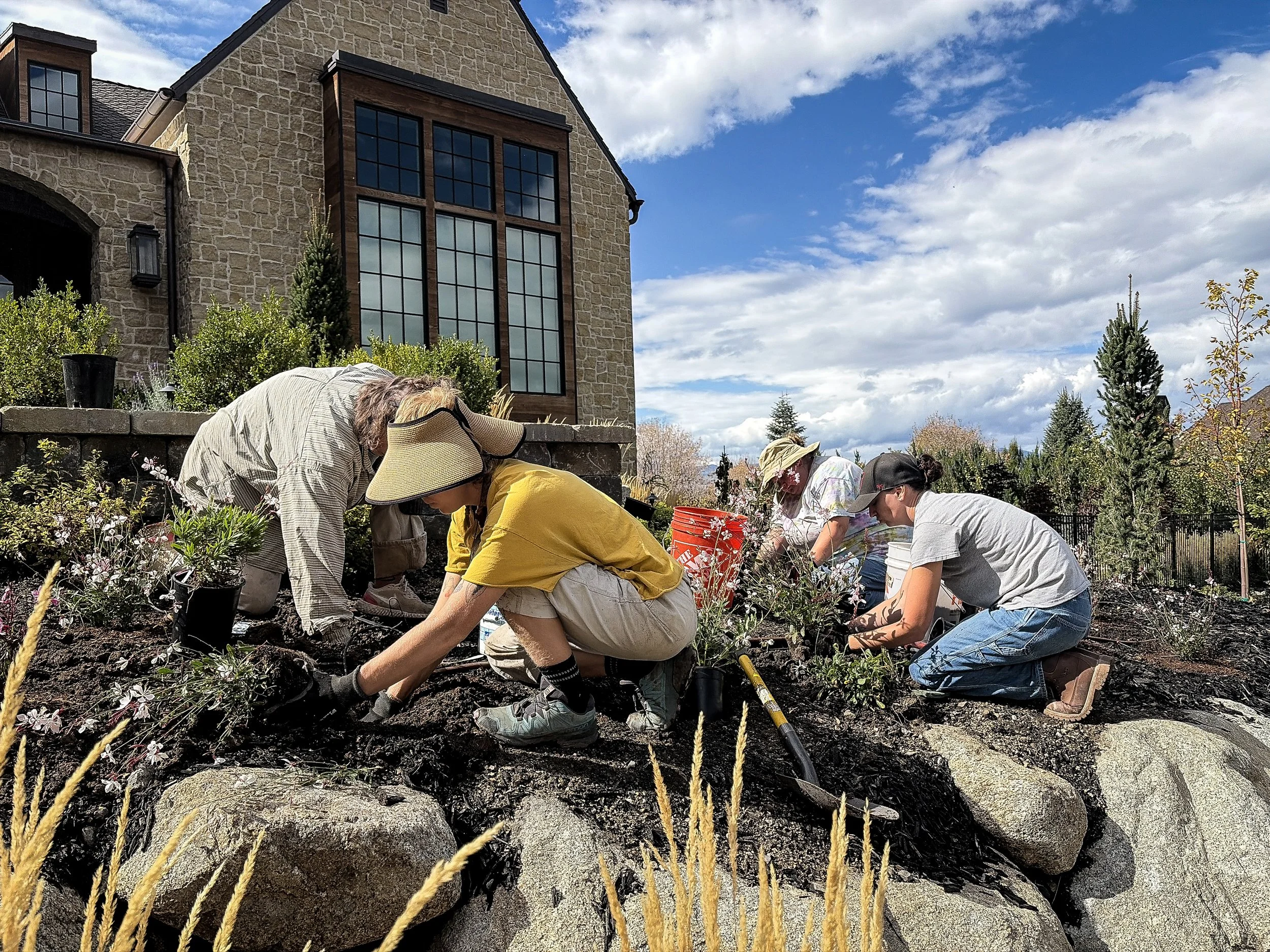 People planting flowers in a garden in front of a house with large windows and stone and wooden siding, under a blue sky with clouds.