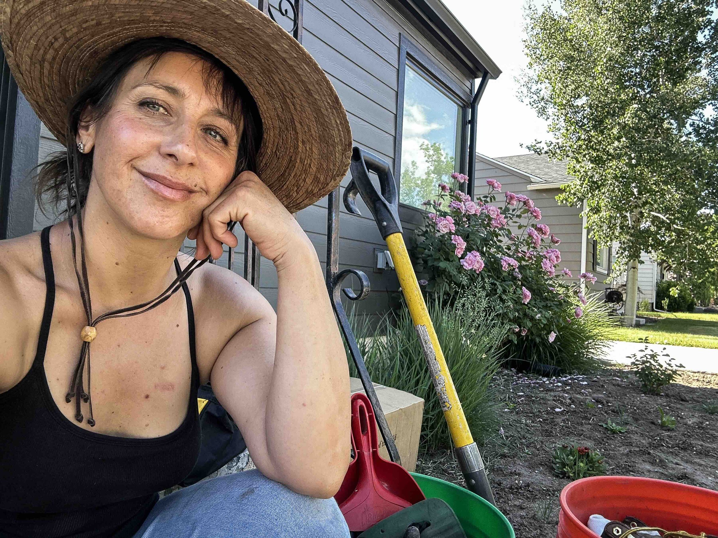 A woman with a wide-brimmed hat and black tank top sitting outdoors, smiling and resting her chin on her hand, with gardening tools and colorful flower pots nearby, in front of a house with a window, trees, and blooming pink flowers.