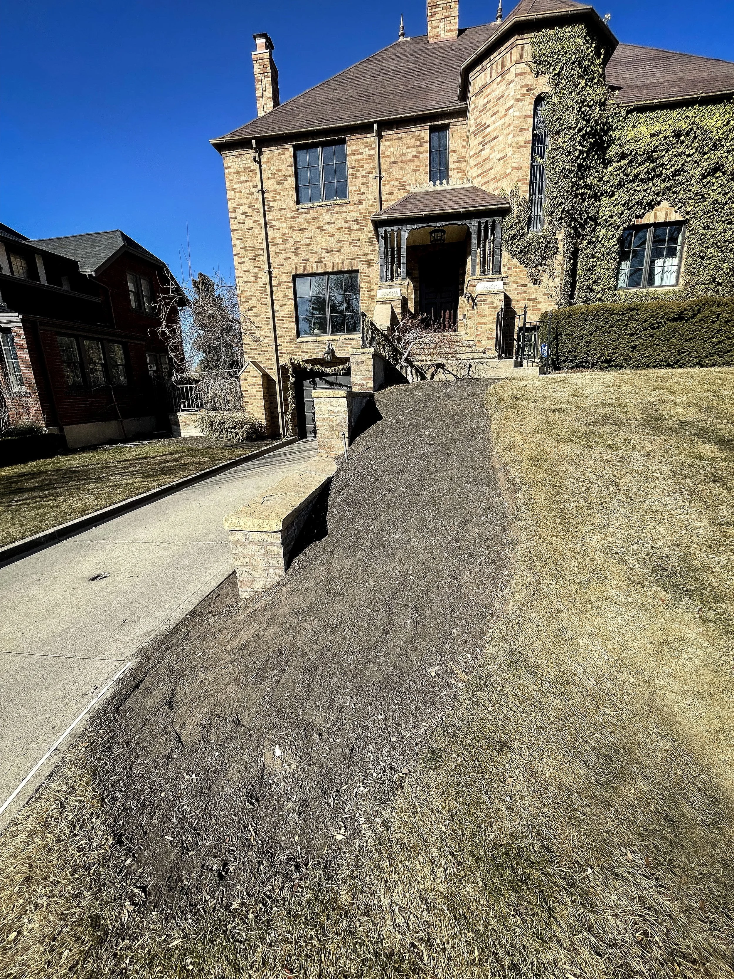 A large brick house with multiple chimneys, black window frames and a front porch, situated on a grassy yard with a sidewalk and a driveway leading up to a garage, under a clear blue sky.