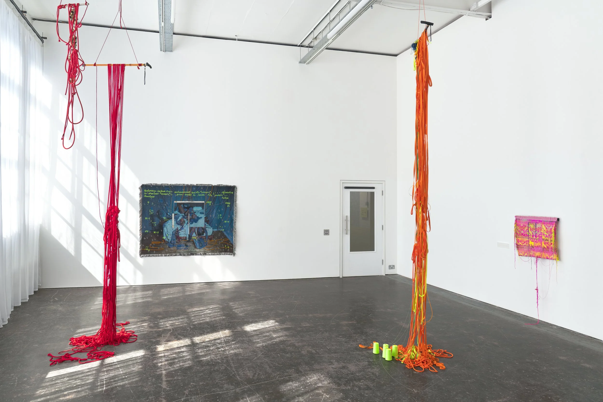 Installation view of Queer Texture at Primary: red and orange textile works suspended from ceiling to floor in a white gallery, with woven pieces on the walls. Photo: Reece Straw.