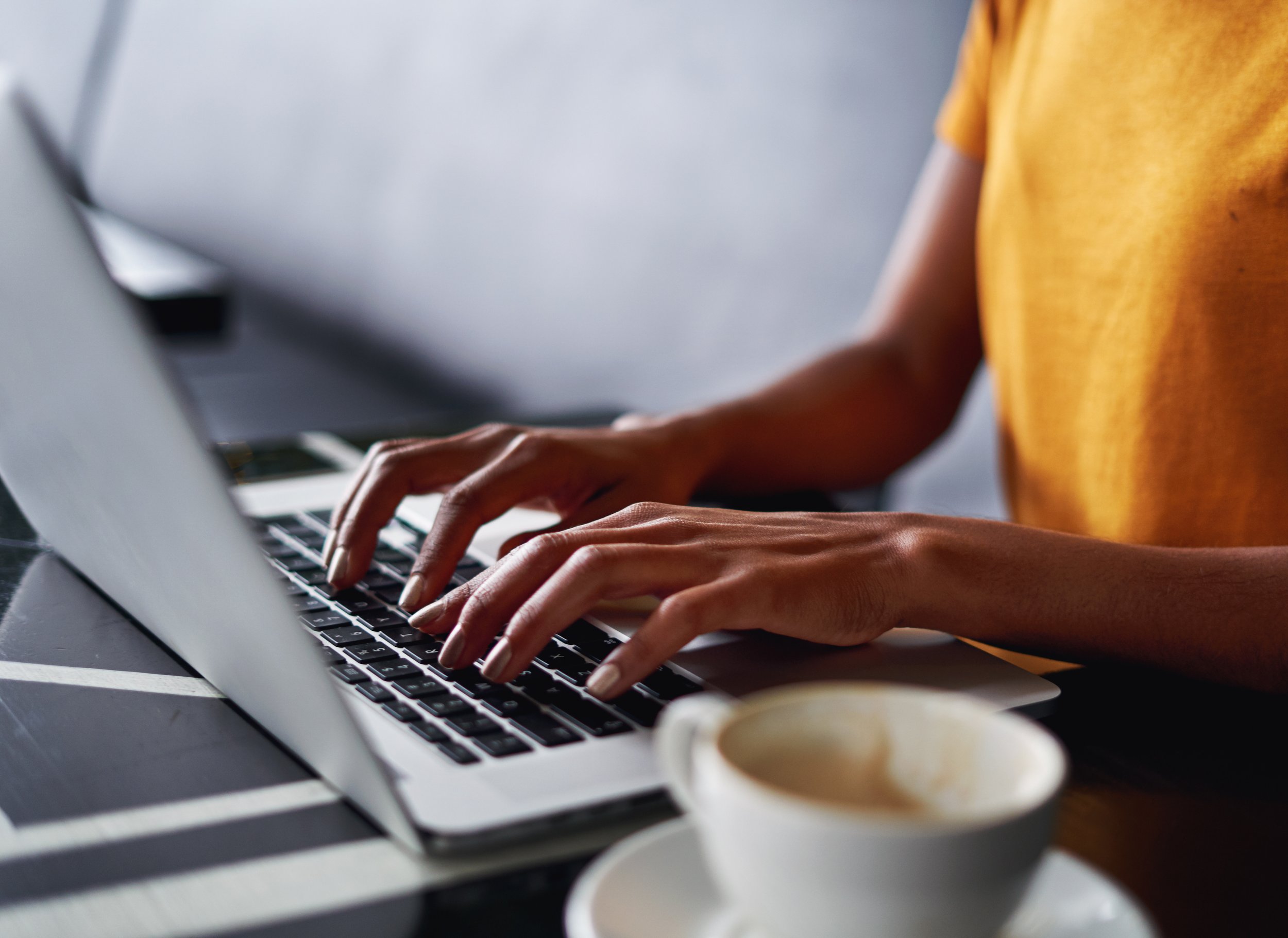 Close-up of a Black person's hands typing on a laptop keyboard, with a white cup of coffee beside it.