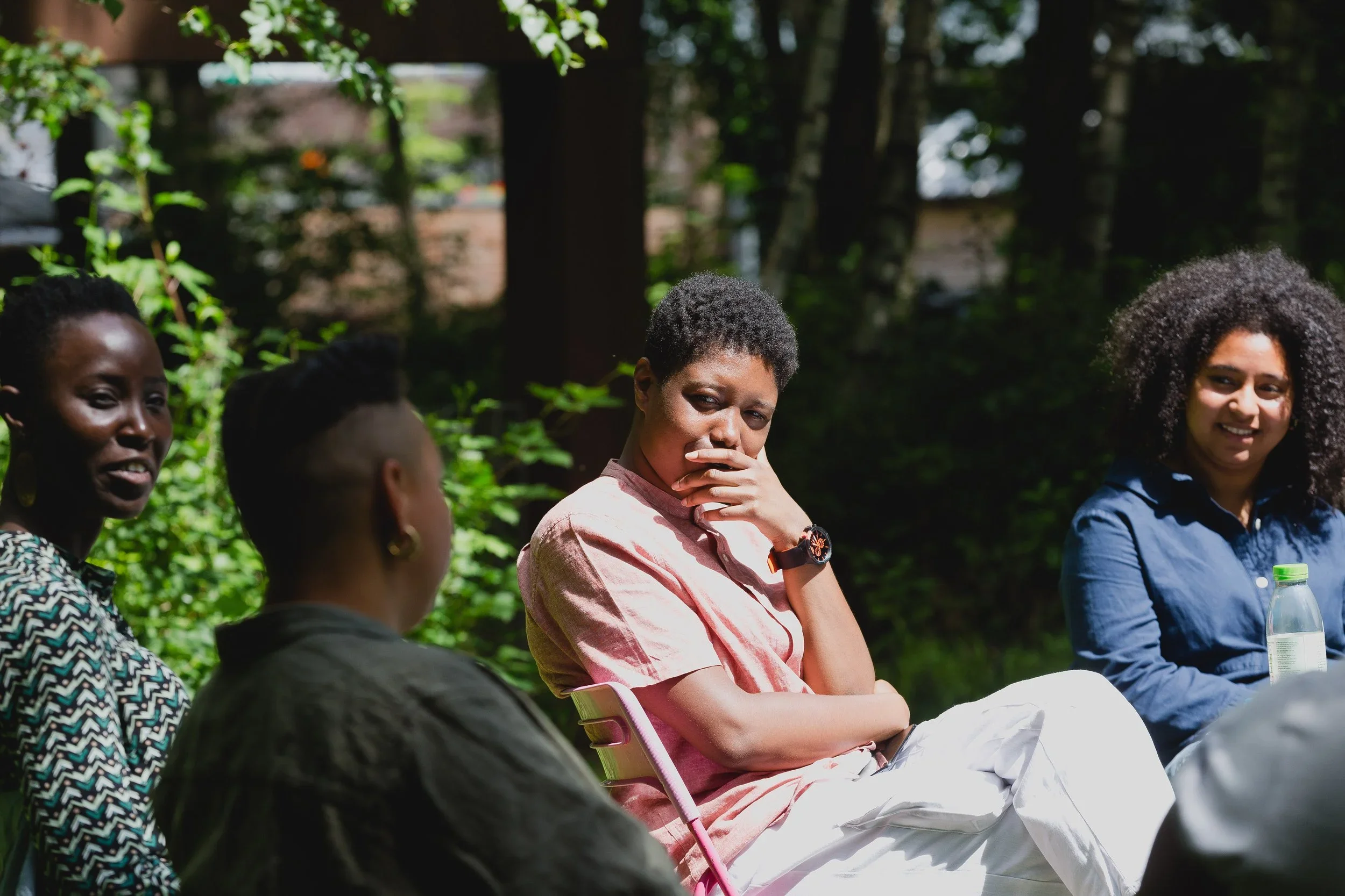 A Black person with short cropped hair, wearing a pink linen dress, sits outdoors in dappled sunlight, hand resting against their chin, listening to someone in the foreground.