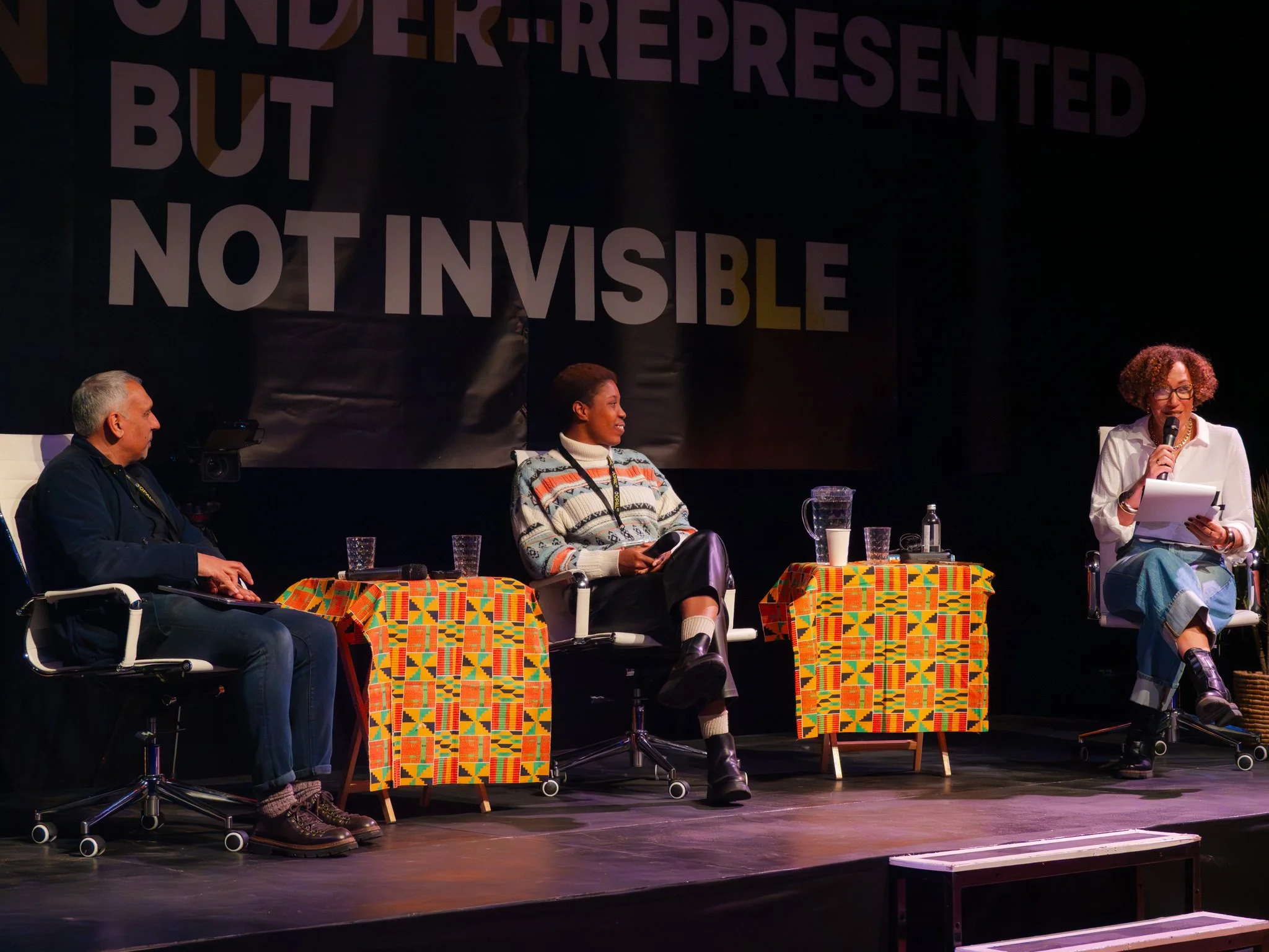 A speaker seated on stage during a panel discussion, with side tables draped in kente cloth, in front of a large banner reading 'Represented, not invisible'.