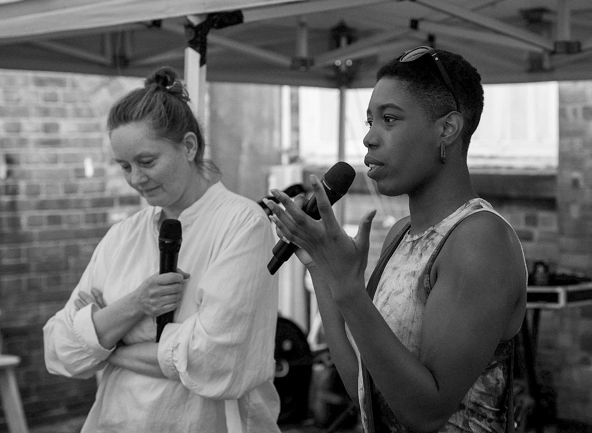 Jade speaking into a handheld microphone, mid-gesture, at an event.