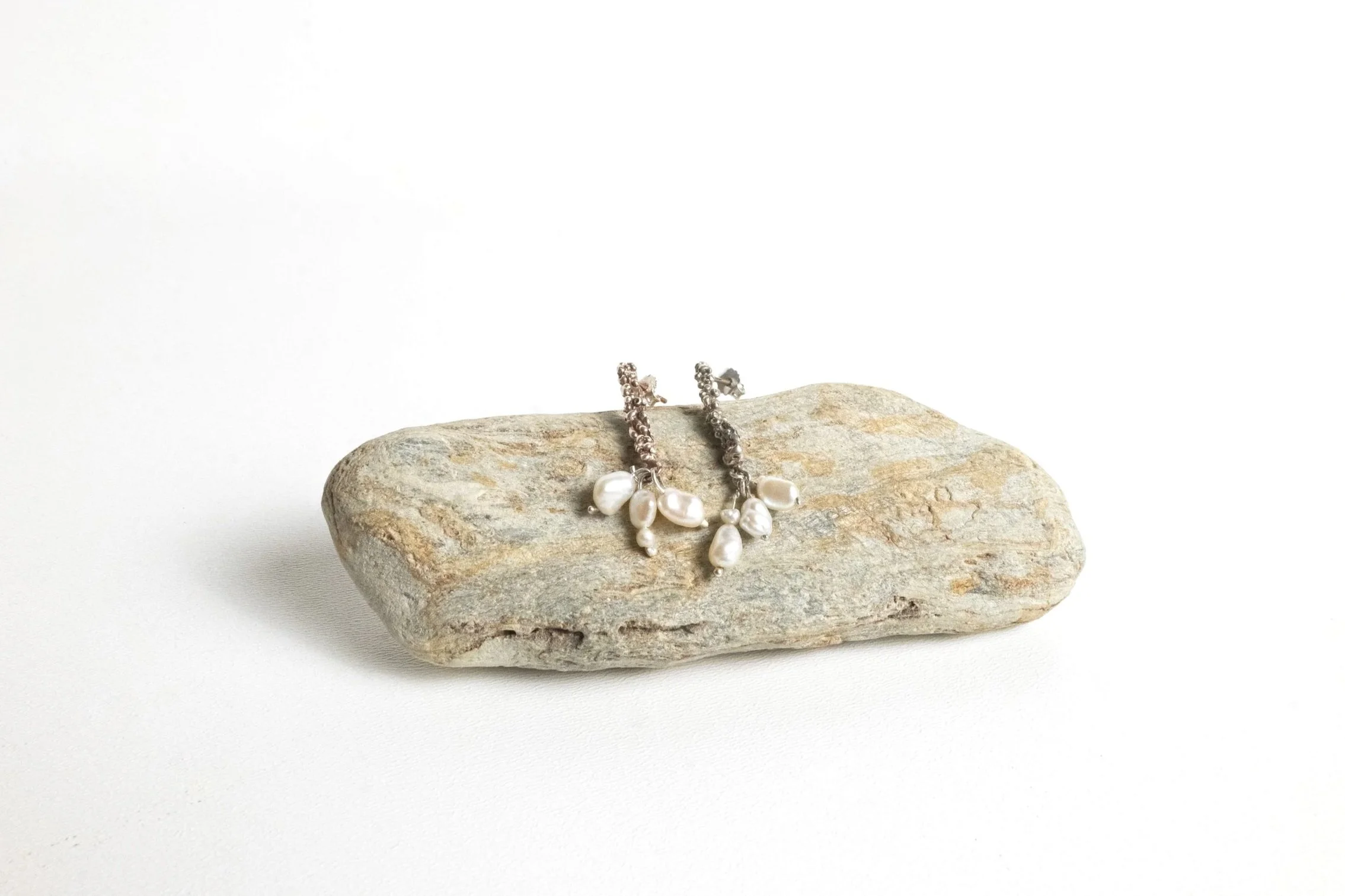 Silver earrings with white pearls resting on a textured rock against a white background.
