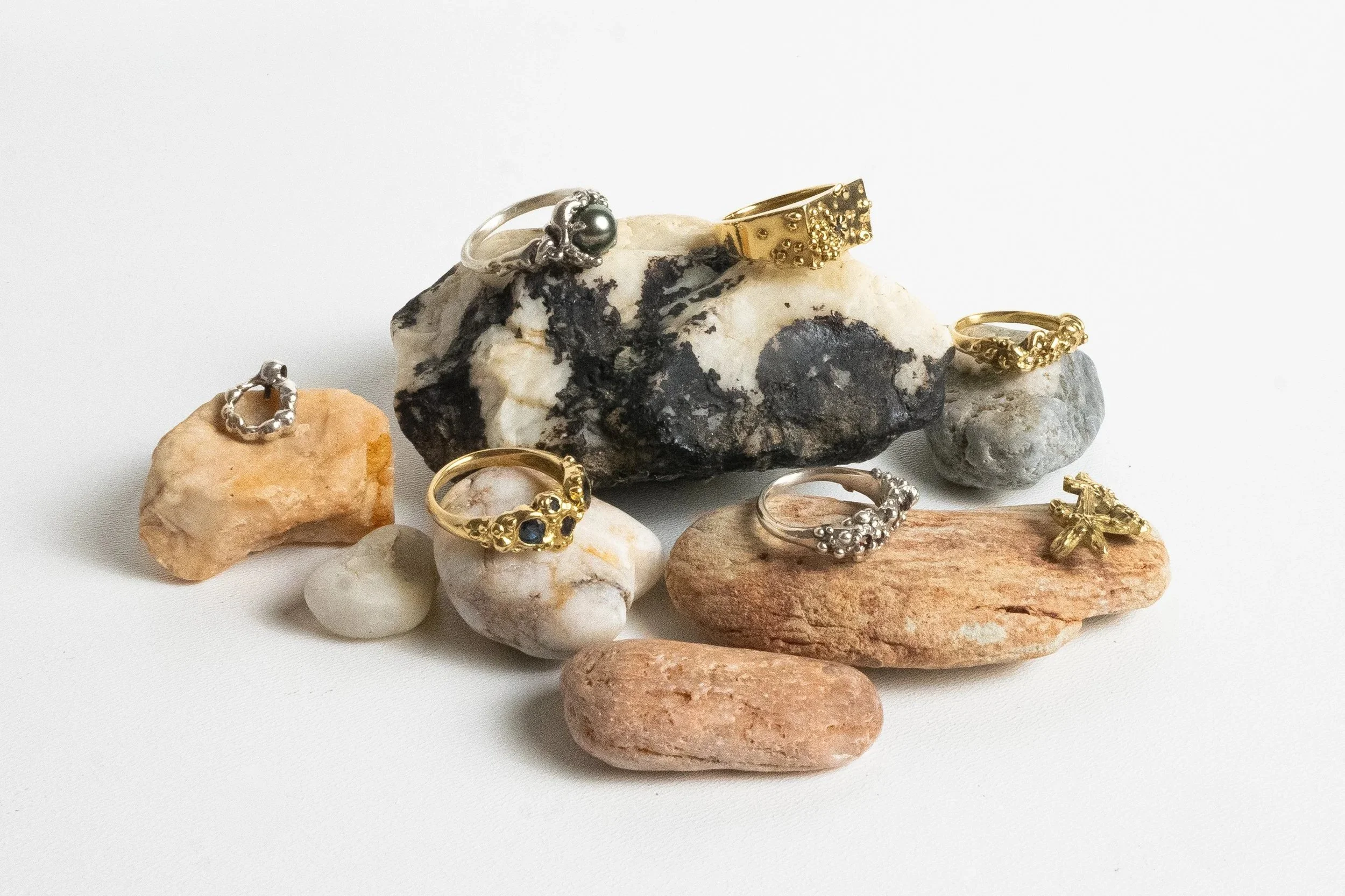 Collection de bijoux composée de bagues en or et en argent, placés sur des pierres naturelles de différentes couleurs et textures, avec un fond blanc.