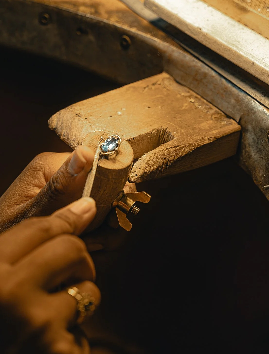 Un artisan en train de fabriquer une bague en or avec une pierre précieuse bleue, sur un support en bois, à l'aide d'outils de bijouterie.
