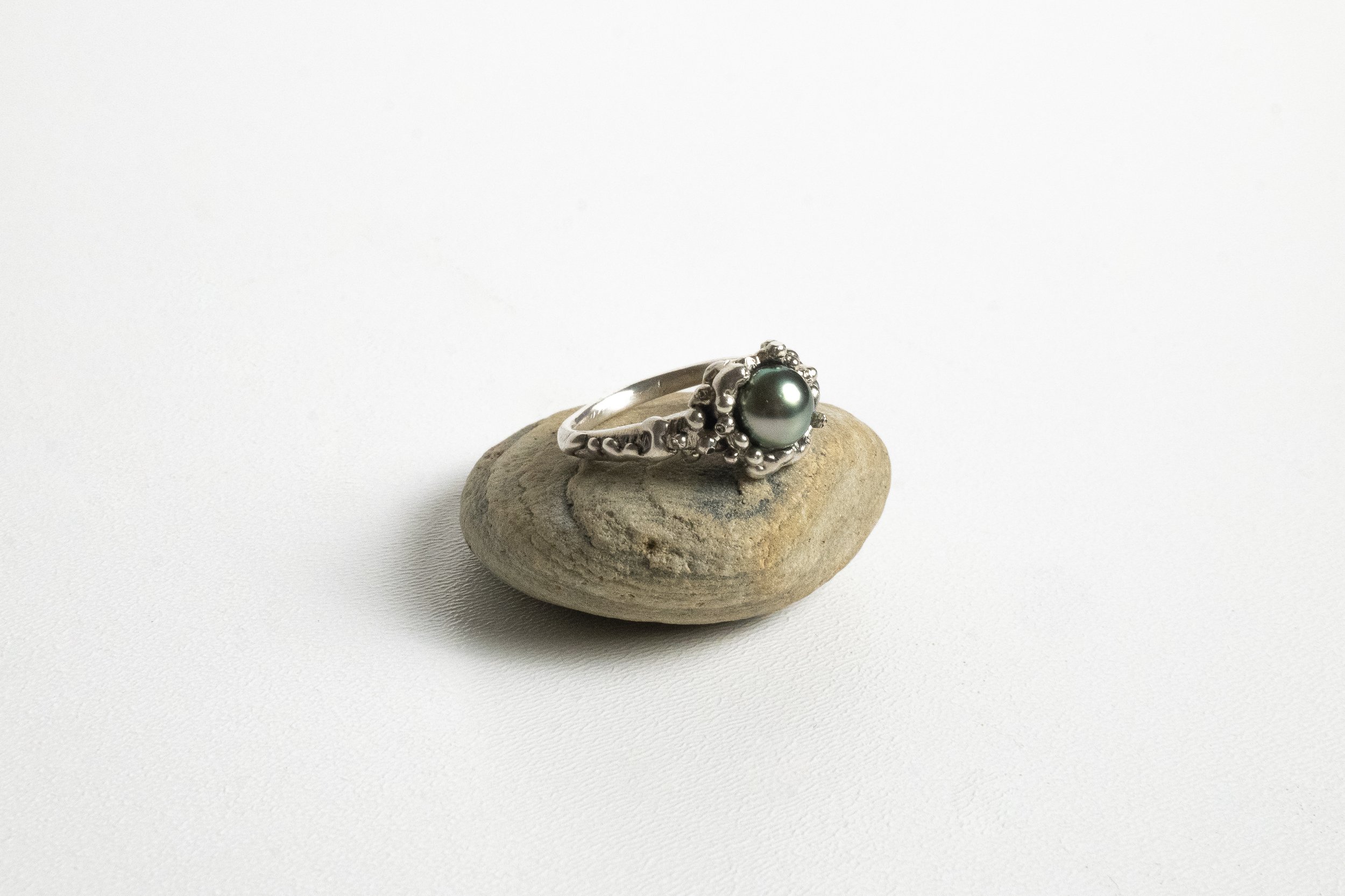 A silver ring with a dark gray pearl in the center, resting on a small, smooth, light-colored stone on a white background.