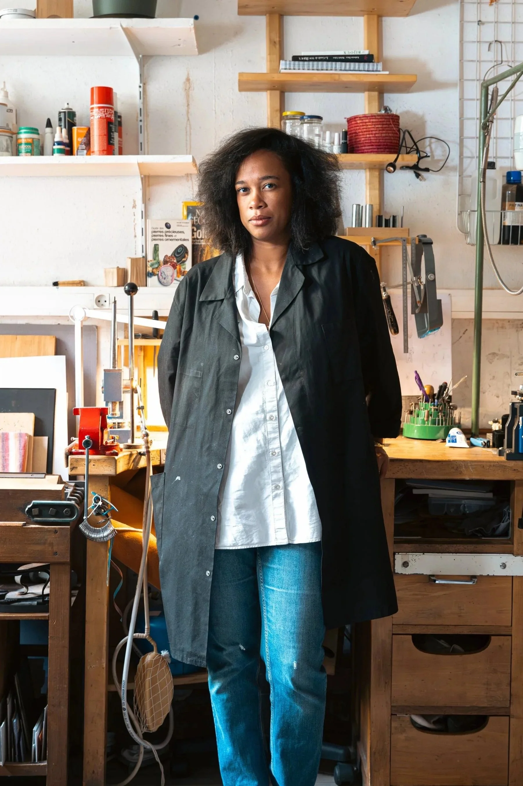 Une femme debout dans un atelier avec des étagères en bois derrière elle, portant une veste noire et un chemisier blanc, entourée d'outils et de matériaux artisanaux.
