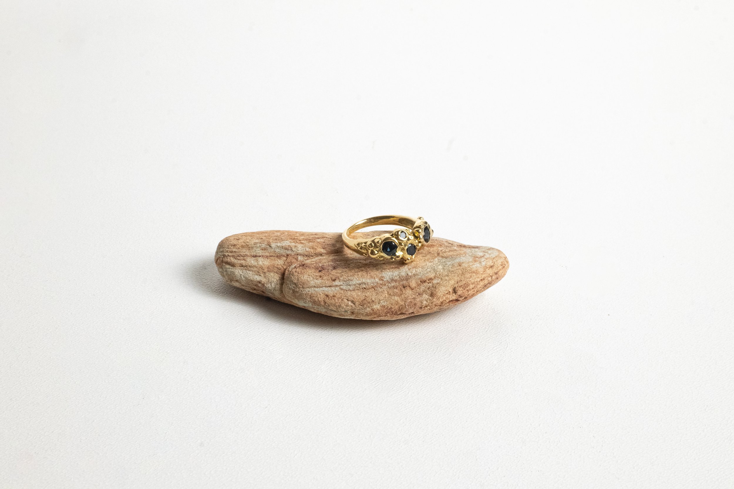Gold ring with black gemstones on a textured brown stone against a plain white background.