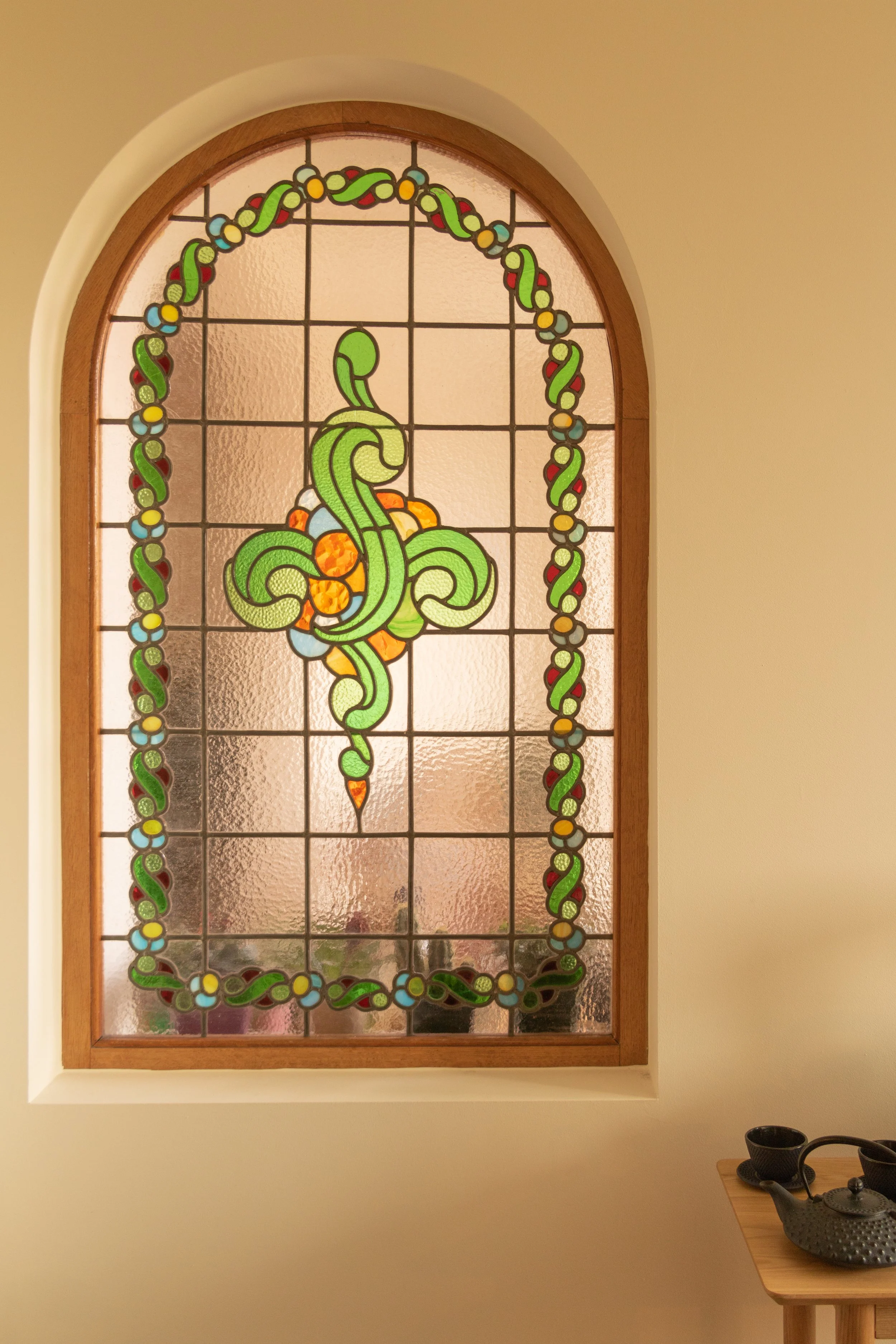 Kleurrijk glas-in-lood raam met een sierlijk, groen patroon en een omlijsting van groene en blauwe kralen, hangend in een boogframe. Aan de onderkant rechts een tafel met een theepot en kopjes.