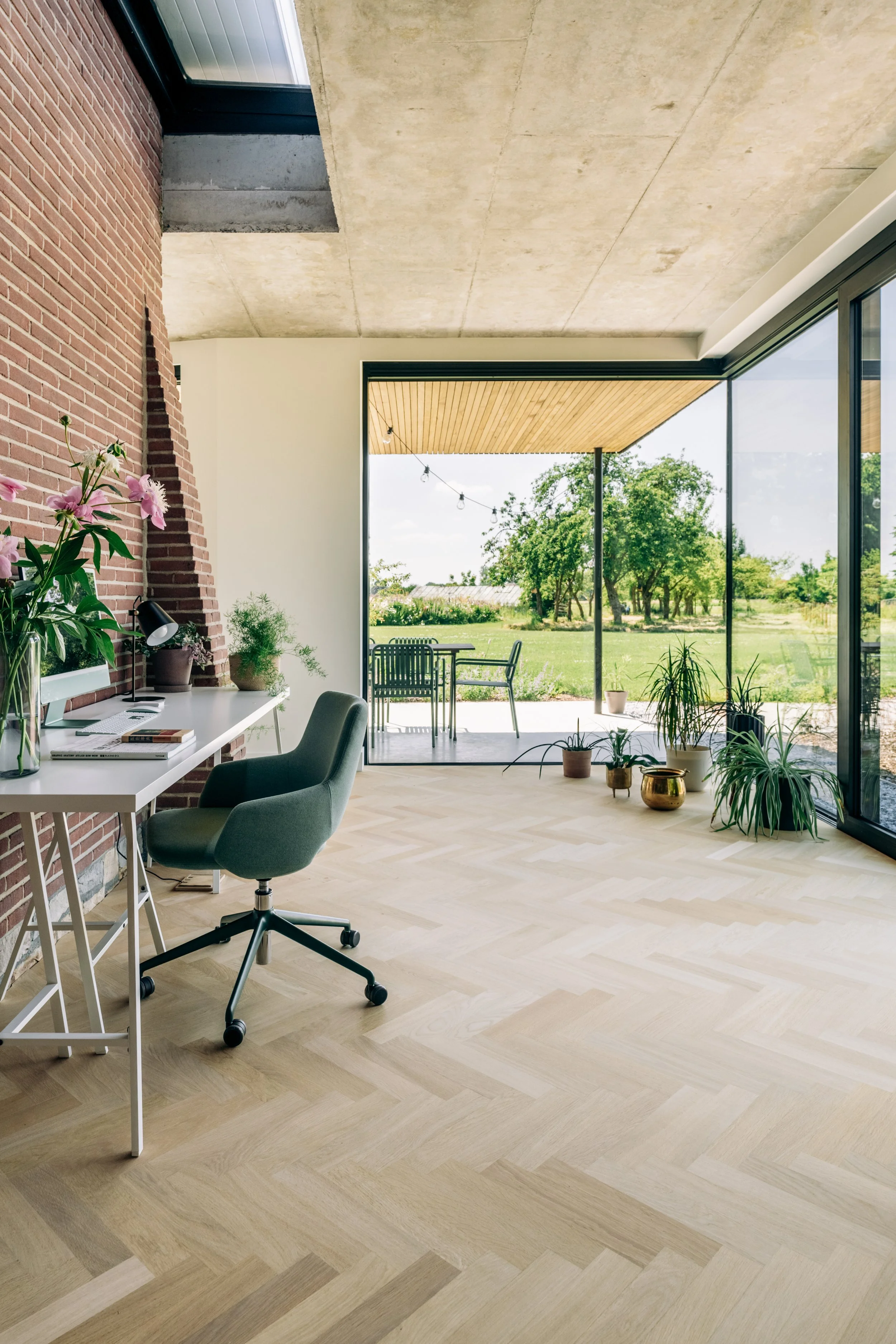 Een modern kantoor of thuiswerkruimte met een witte bureau, een groene stoel, planten, en een grote glazen muur die naar een tuin leidt met bomen en een terras met meubels.