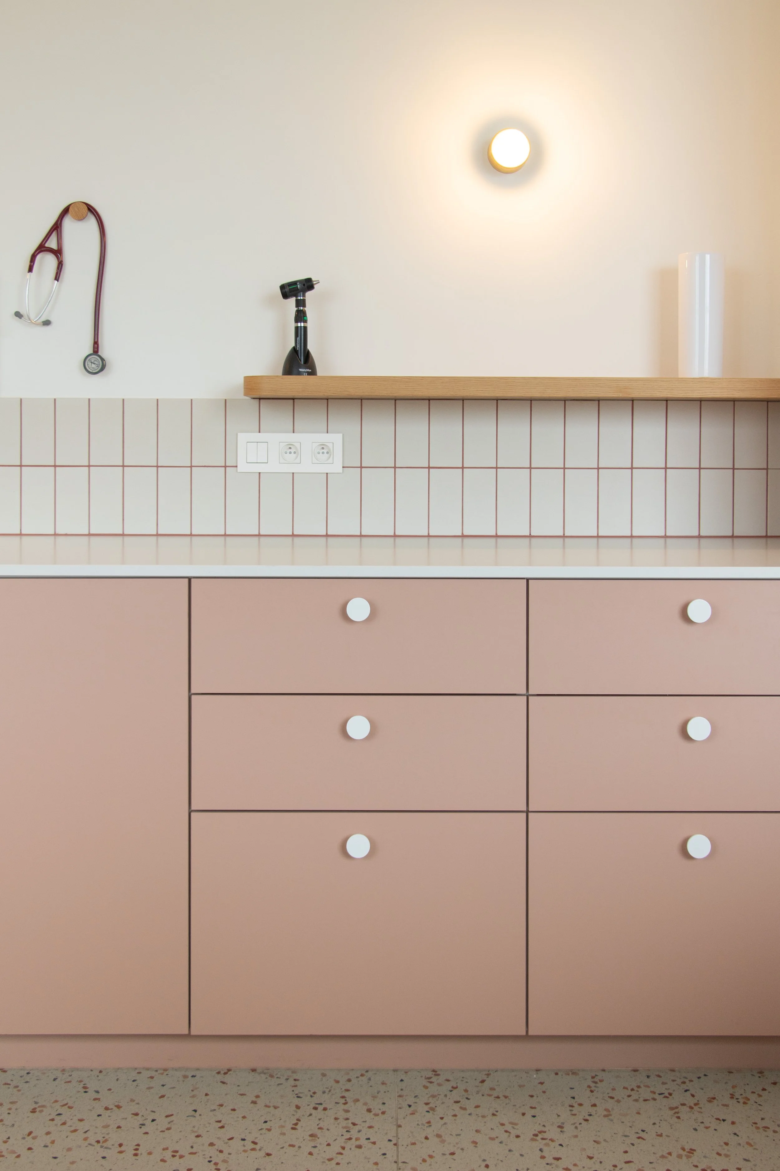 Een kitchenette met een lichtroze kast, witte knopdeuren, een werkblad, een stopcontact, een stethoscoop, een spuitpistool en een witte pilaar, met een muurverlichting en rode betegeling.