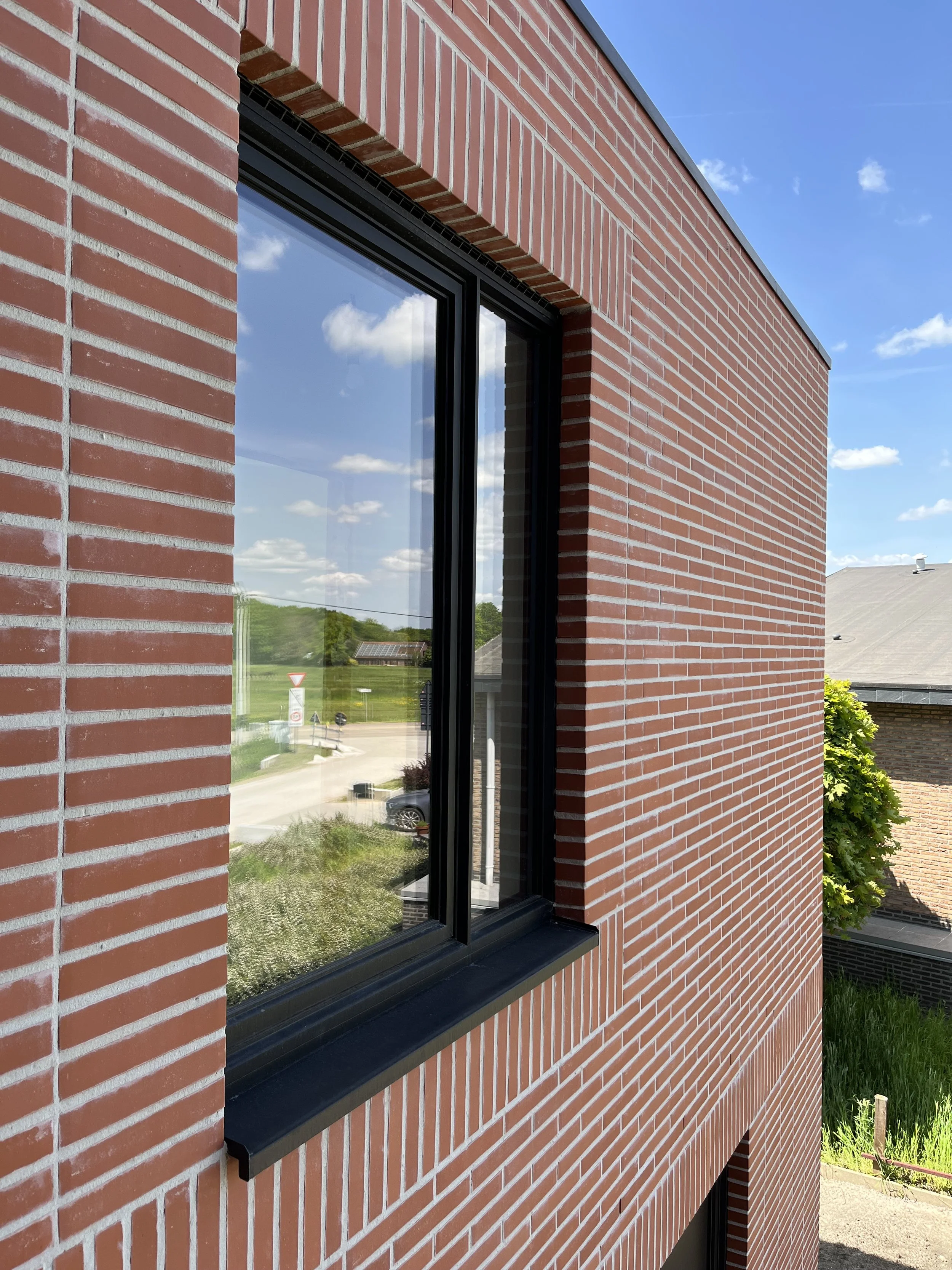 Close-up van een rood bakstenen gebouw met een grote raam met zwarte kozijnen, weerspiegelt de blauwe lucht en witte wolken.