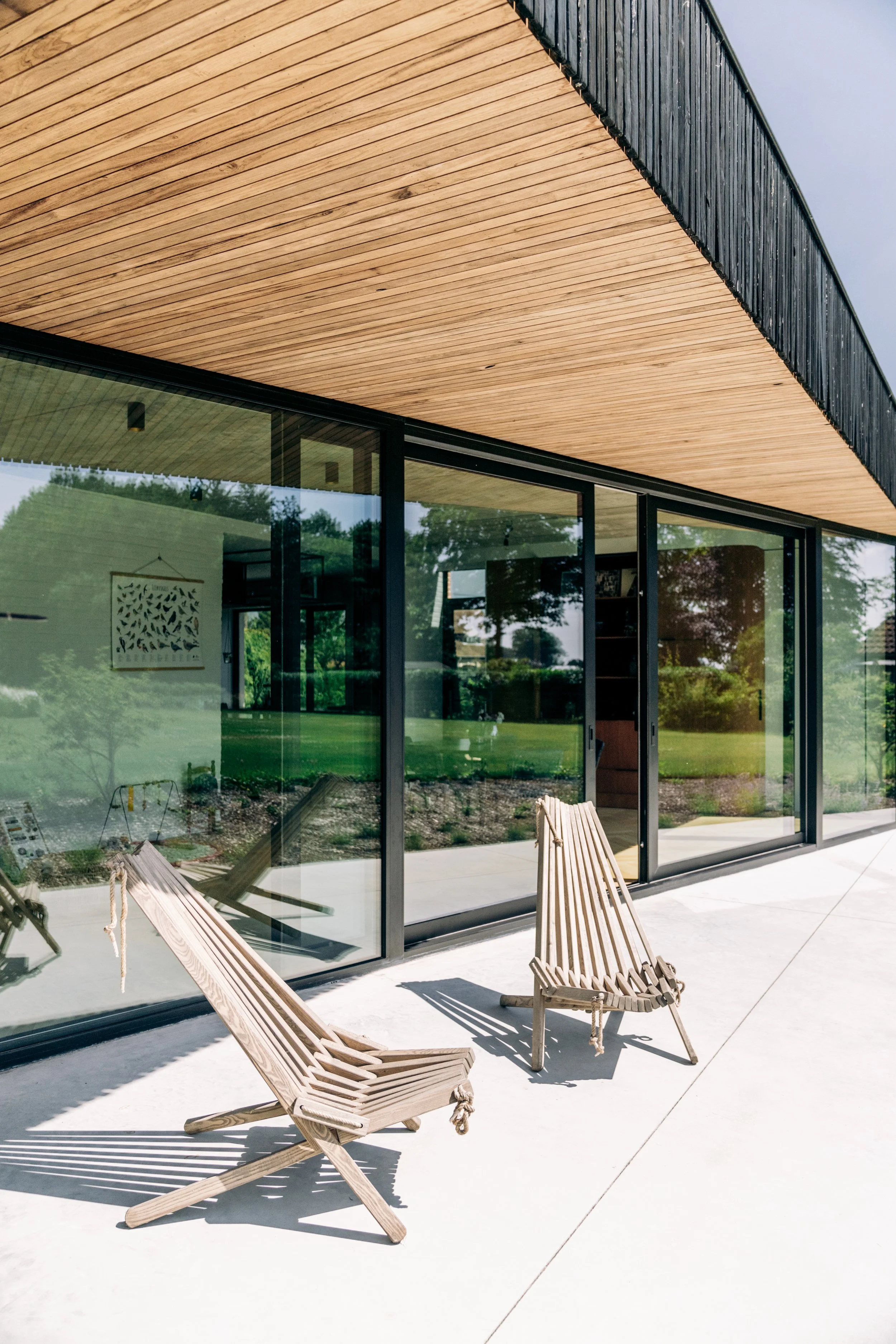 Twee houten loungestoelen op een patio voor een modern huis met grote glazen deuren en een houten plafond.