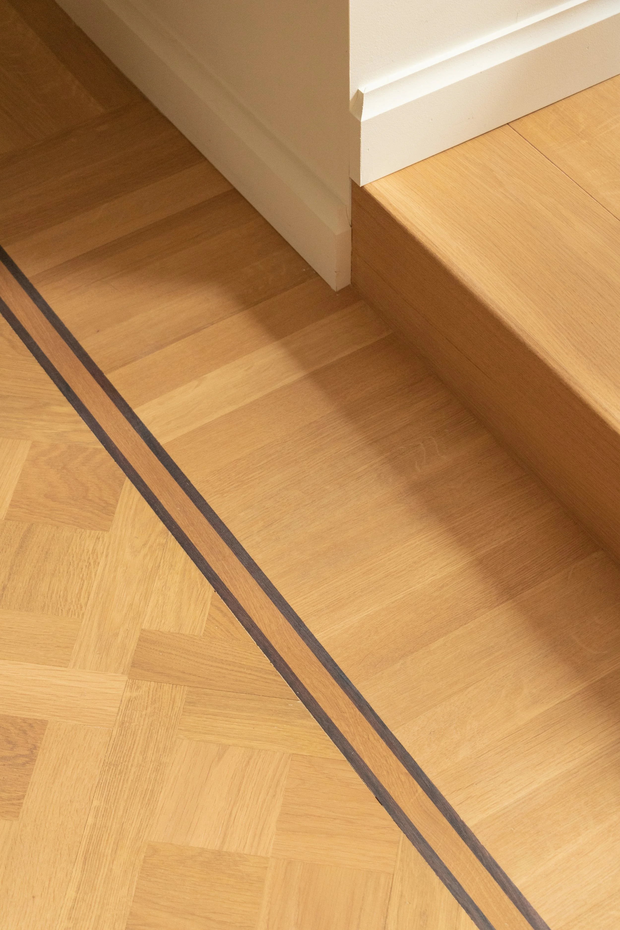 De afbeelding toont een hoek van een houten vloer en een trap, met een witte muur en witte basisplint.