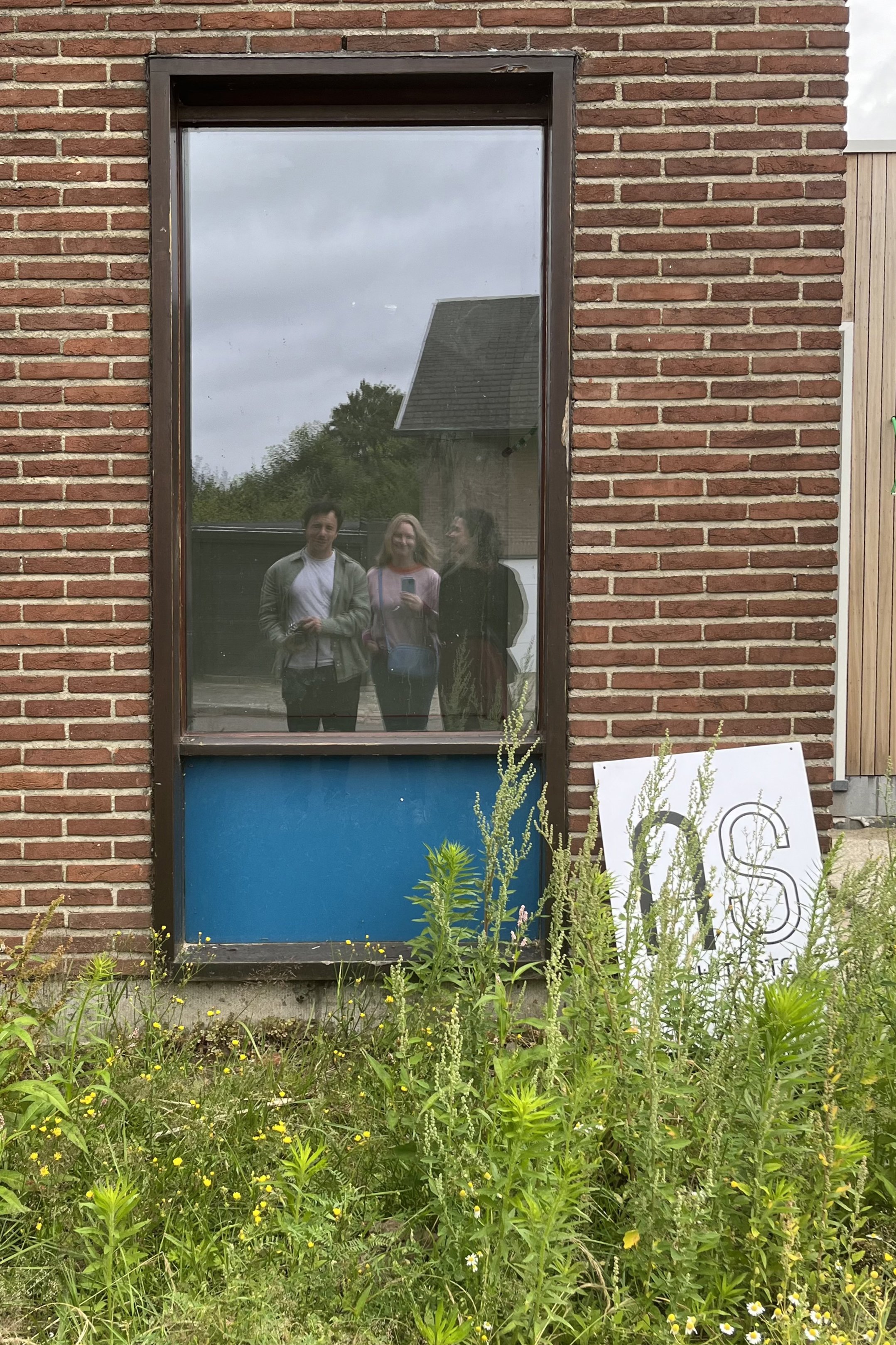 Reflectie van drie mensen in een raam van een rood bakstenen gebouw, met planten en een bord met letters op de grond in de tuin voor het raam.