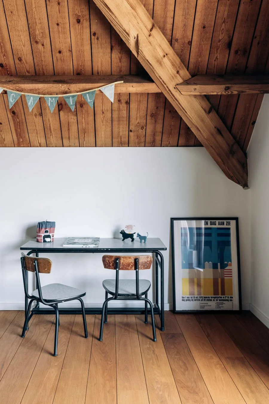 Interieur van een kamer met een houten plafond, een witte muur en een houten vloer. Een klein bureau met twee stoelen, decoratie op het bureau en een poster die op de vloer leunt.