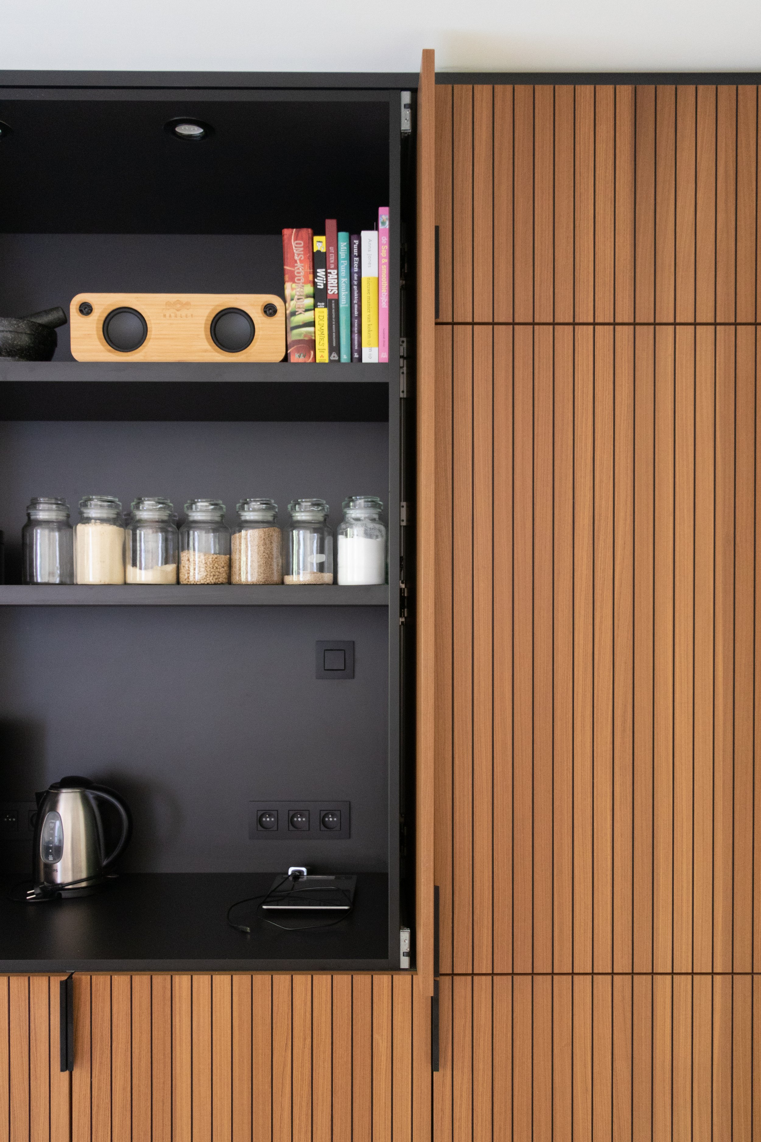 Keukenwand met houten kasten, zwarte planken, een elektrische waterkoker, een broodrooster, glazen potten met ingrediënten, een lade, en een stekker met een apparaat.