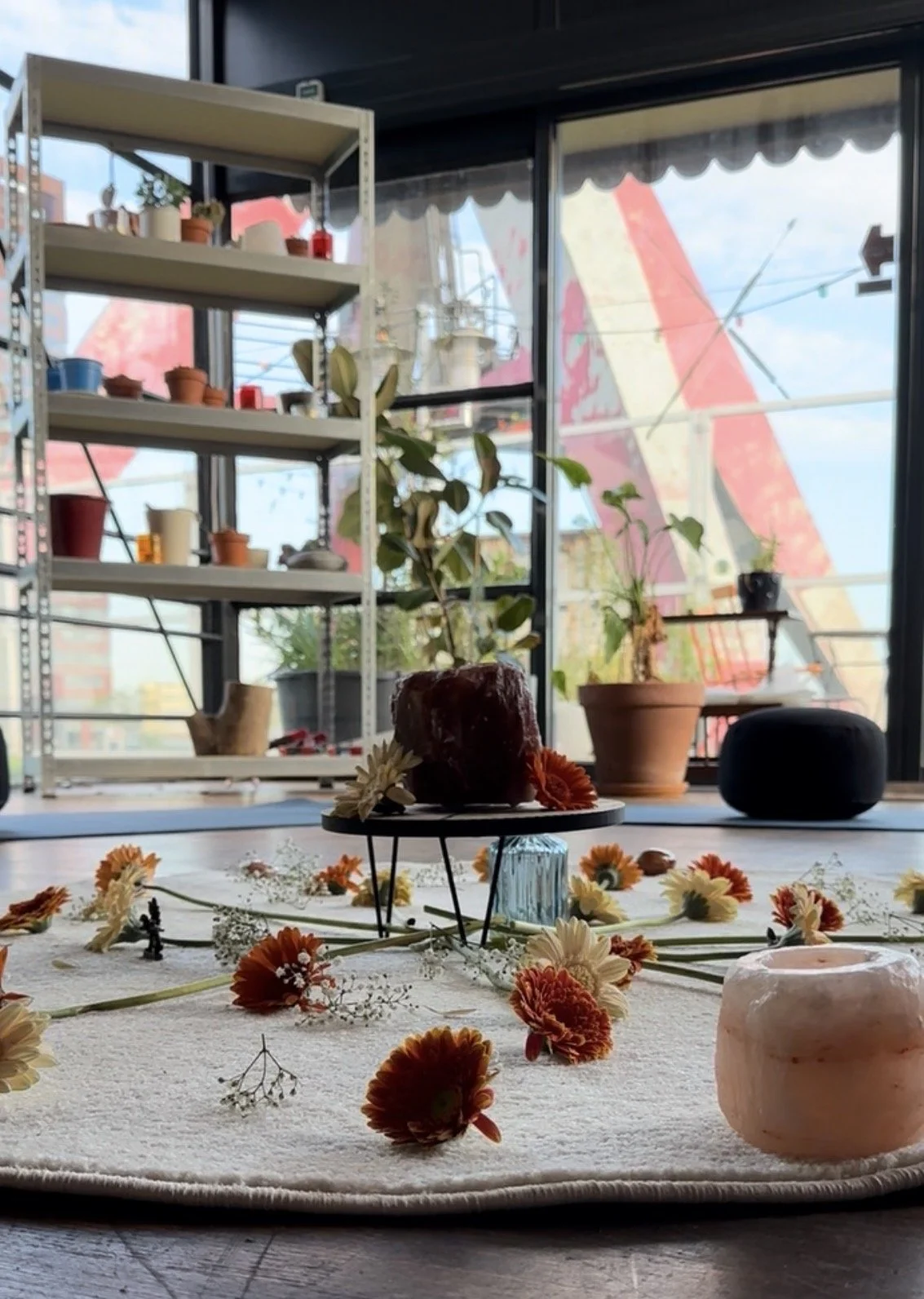 Binnenruimte met planten op de vensterbank en een display met bloemen en decoraties op een tapijt met een grote uitkijk op de stad buiten door grote ramen. Workshop, teambuilding, verbinding, persoonlijke groei, boeddhisme.