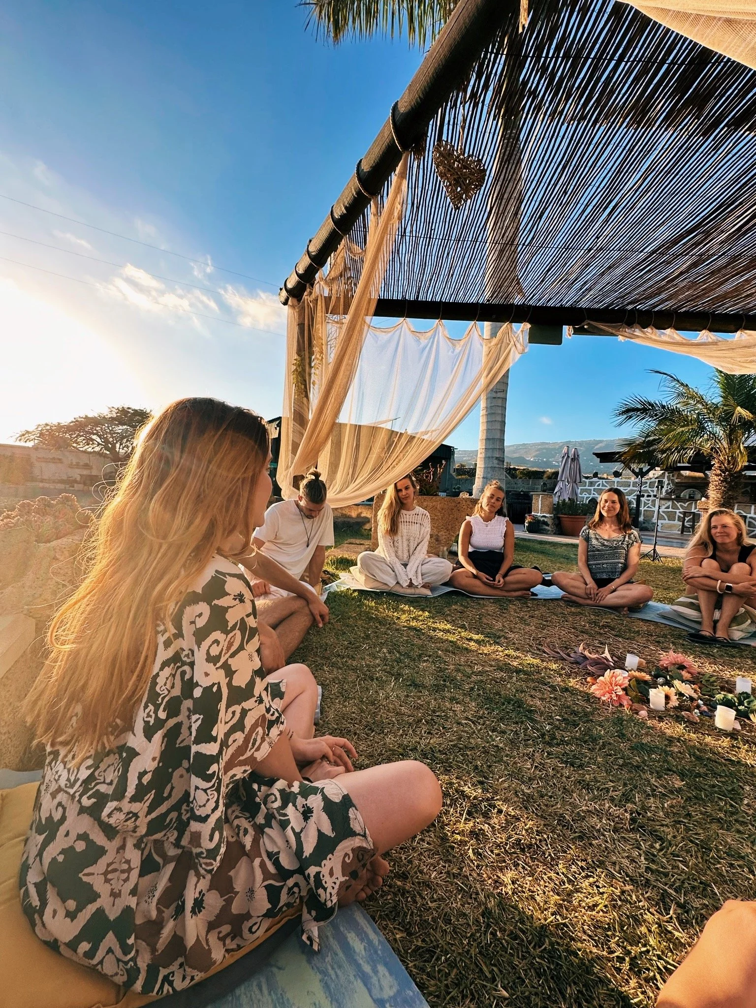 Groep mensen zit in een cirkel op het gras buiten onder een houten en bamboe overkapping met decoraties zoals harten en bloemen, tijdens een bijeenkomst of meditatie, met een heldere blauwe lucht en een landschap op de achtergrond.