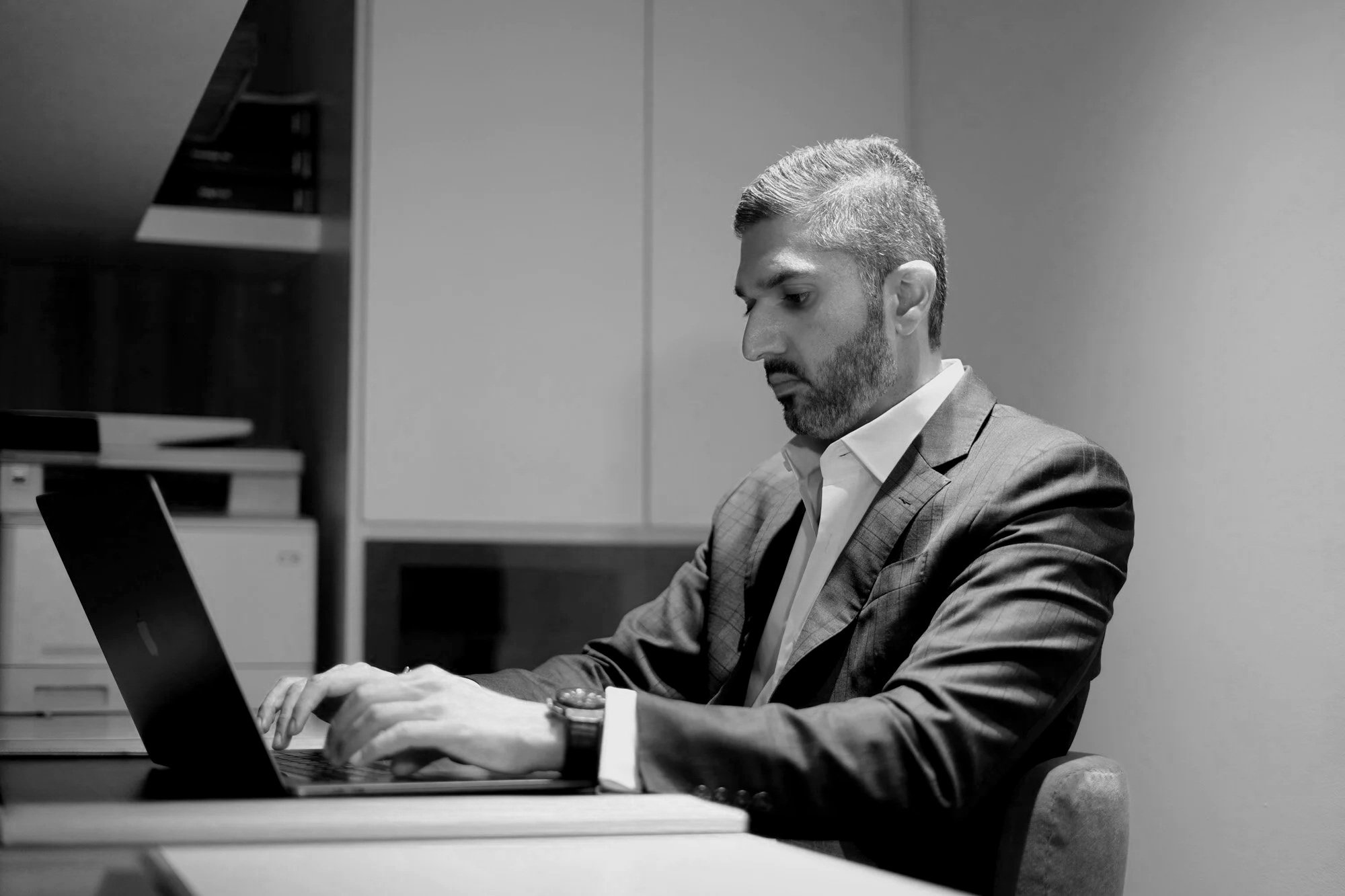 A man in a business suit with short, styled hair and a beard is sitting at a desk, using a laptop. The background shows office furniture including a filing cabinet and shelves.