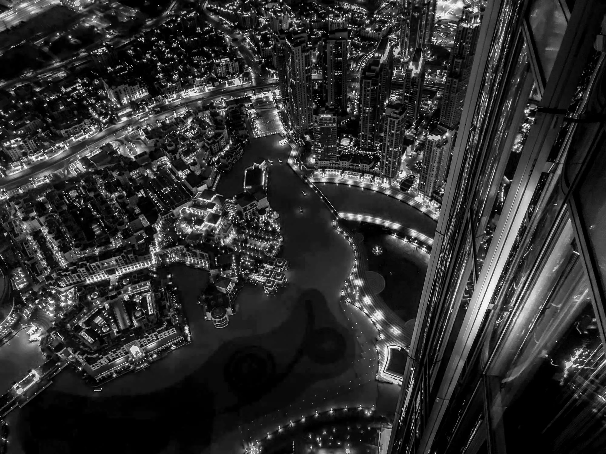 Nighttime cityscape with illuminated roads, buildings, and waterways seen from a high vantage point, featuring tall skyscrapers and urban lights.