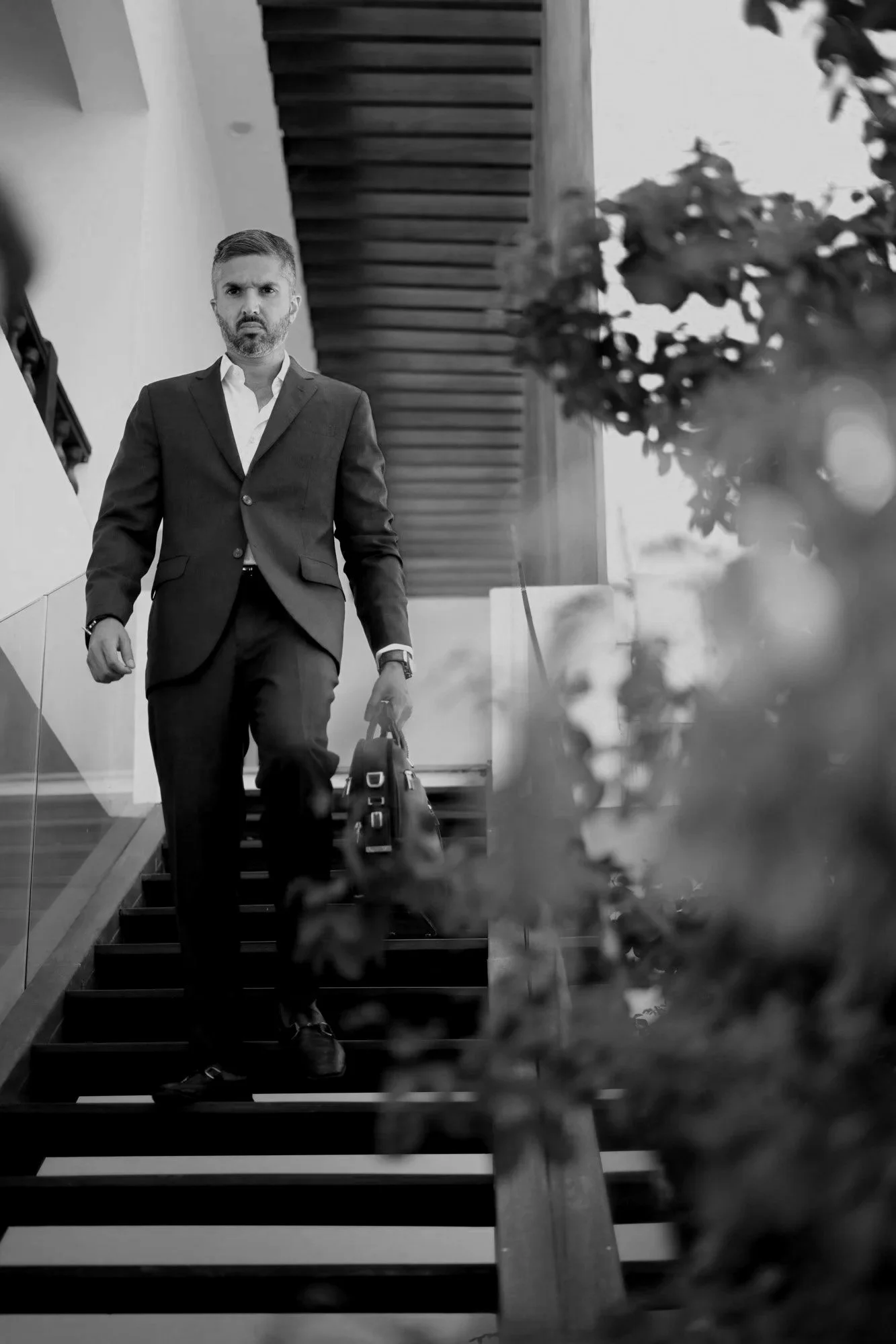 A man in a suit walking up stairs while carrying a briefcase, with plants in the foreground and modern architectural elements in the background.