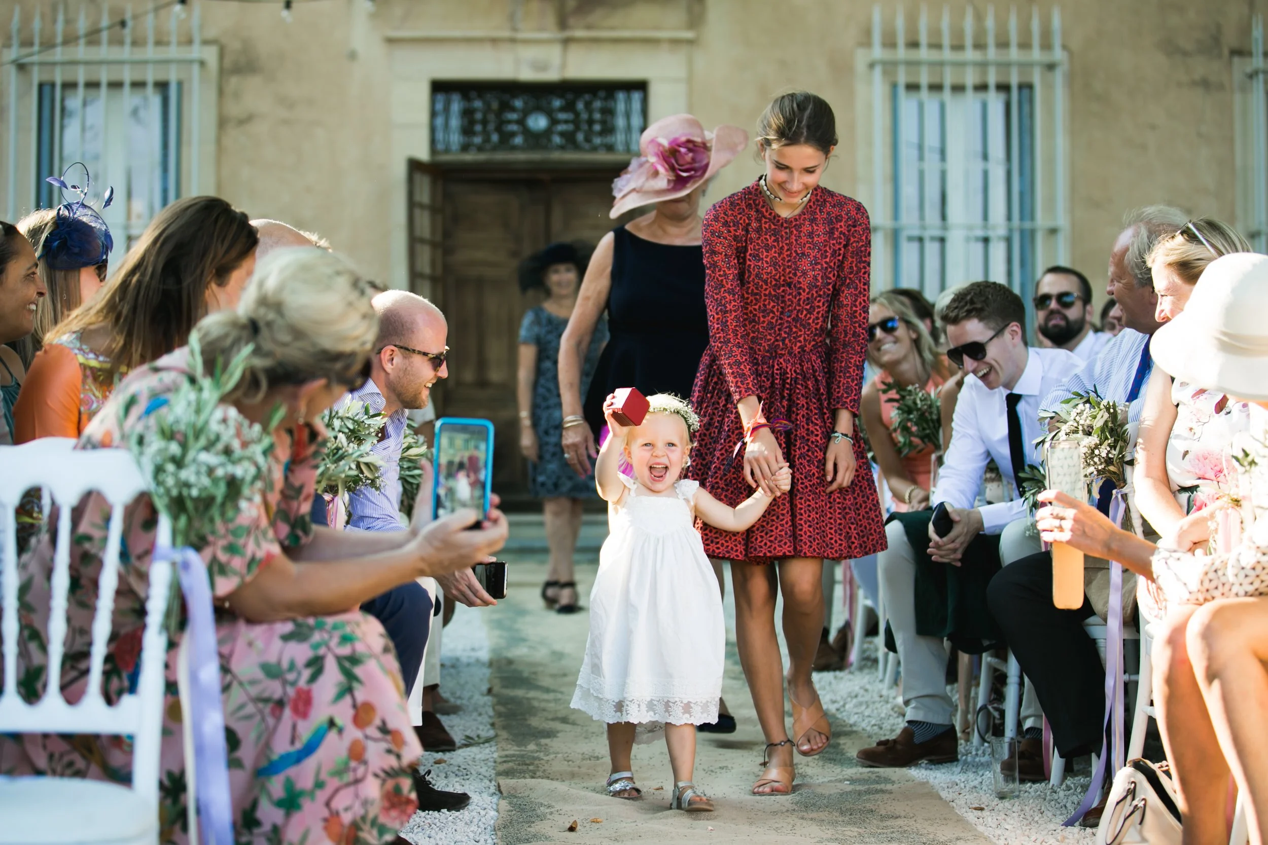 Een bruid en kleine meid lopen hand in hand door een rij gasten tijdens een bruiloft. Gasten zitten en staan langs de zijkanten, sommigen maken foto's en lachen.