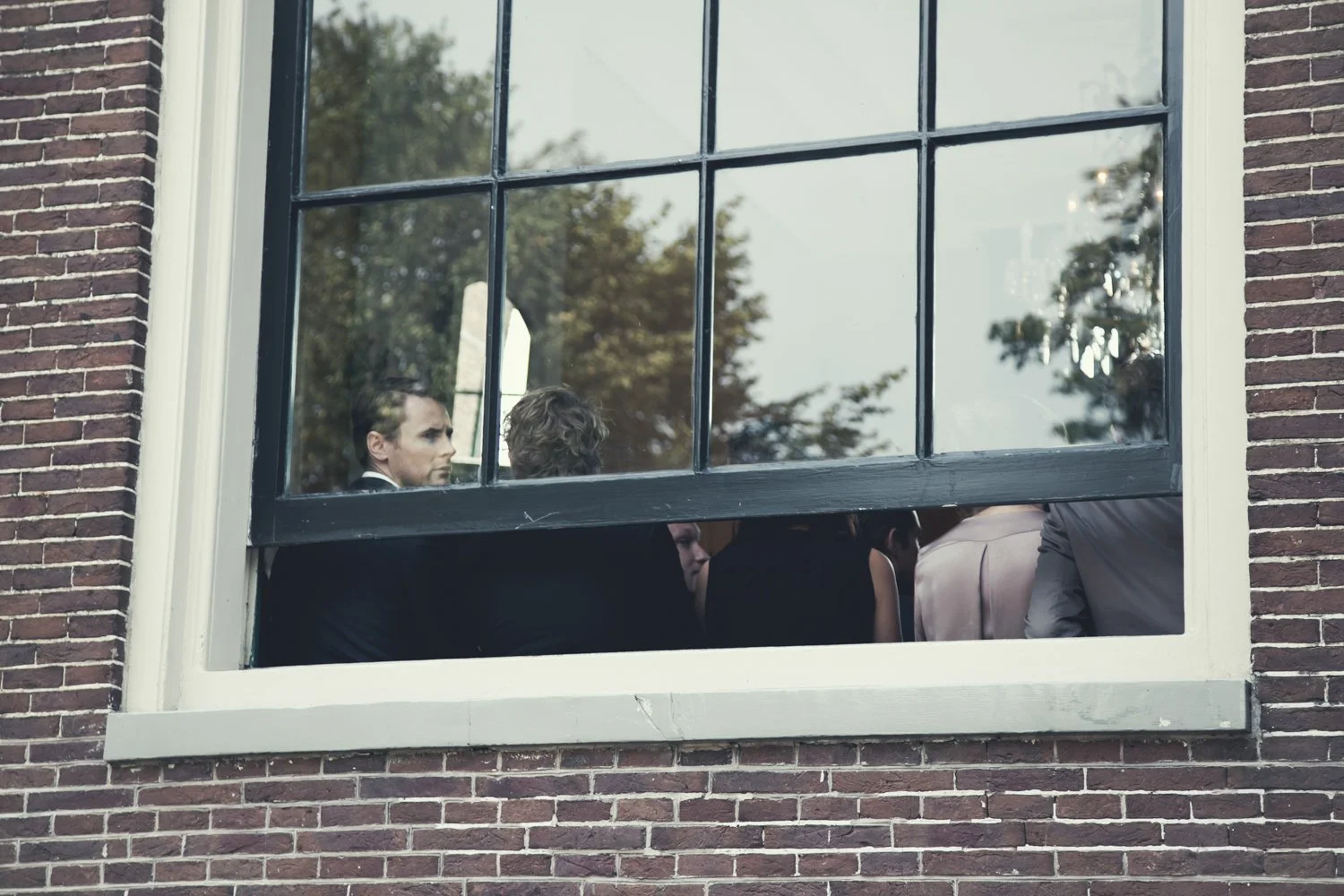 Mensen die binnen in een kamer of hal zitten, te zien door een raam met meerdere vierkante ruiten, afgezien van een gevel met bakstenen muur.