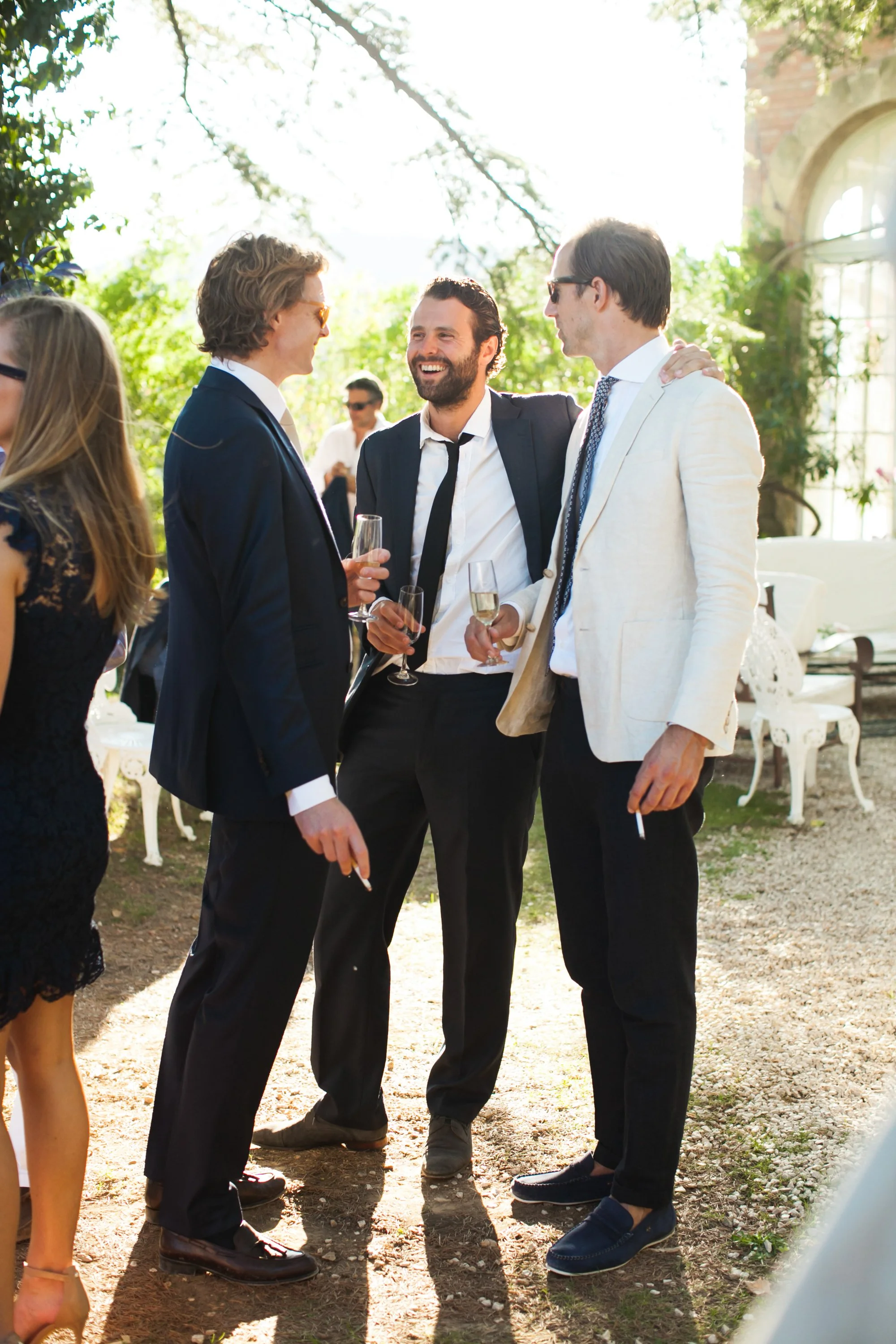 Vijf mensen in formele kleding die samen lachen en praten, terwijl ze wijn drinken op een buitenfeest in de zon.