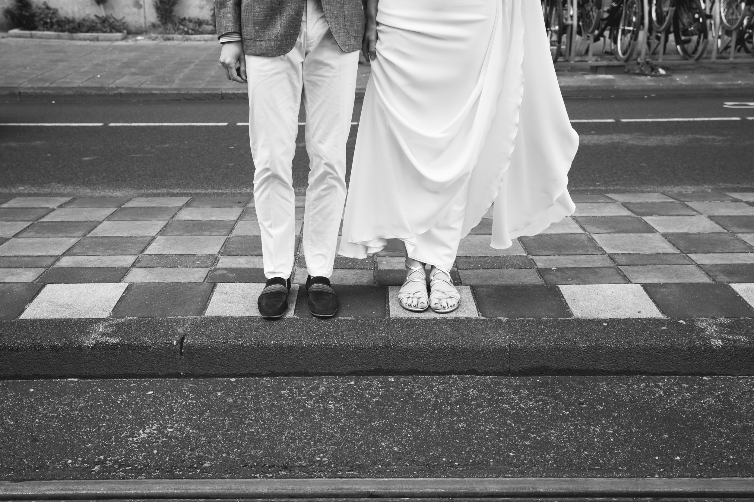 Close-up van de benen en schoenen van een getrouwde paar, staand op een zebrapad in zwart-wit, met de man in een grijze jas en lichte broek en de vrouw in een witte jurk en sandalen.