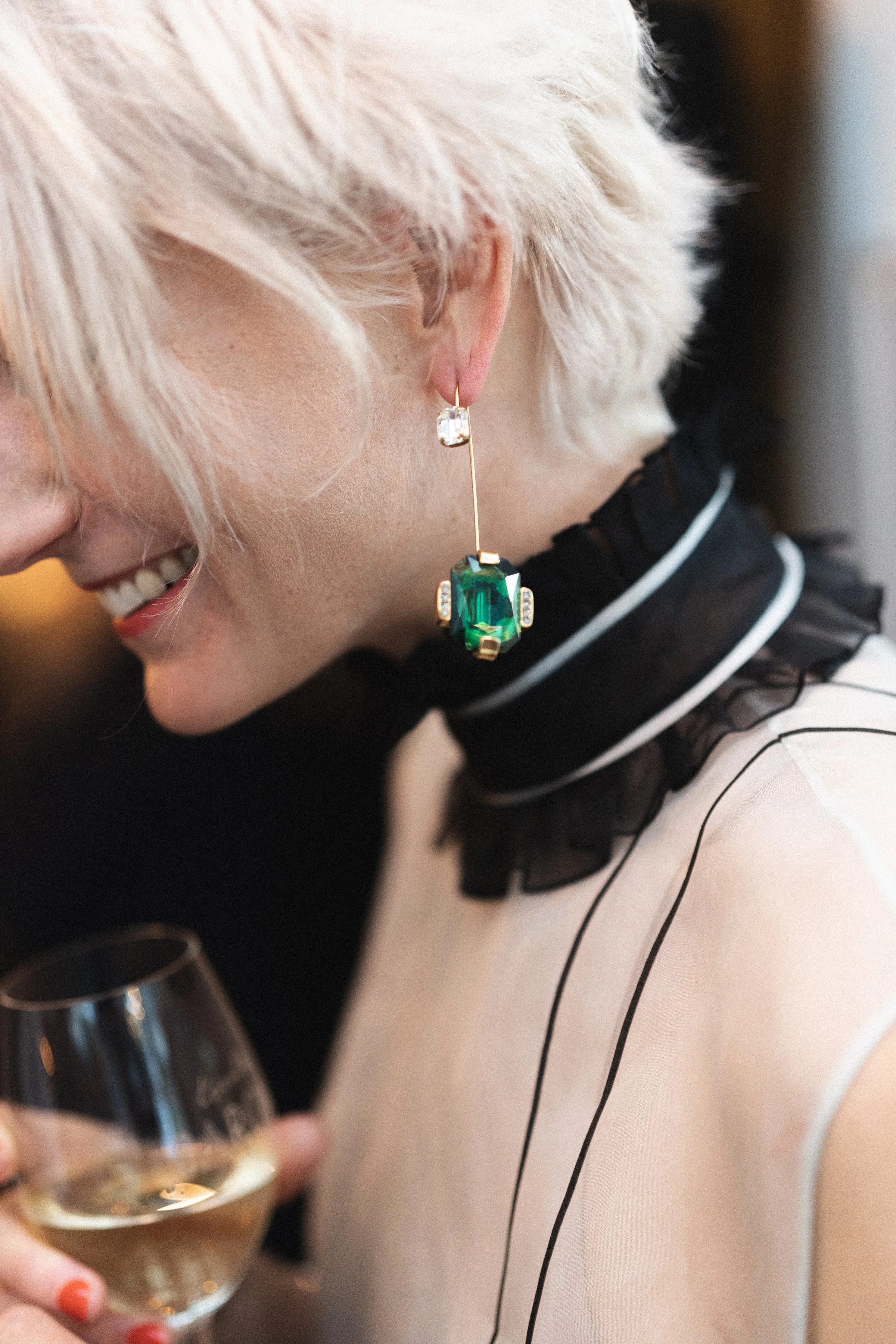 Close-up van een lachende vrouw met een grote groene en diamanten oorbel, die een glas witte wijn vasthoudt.