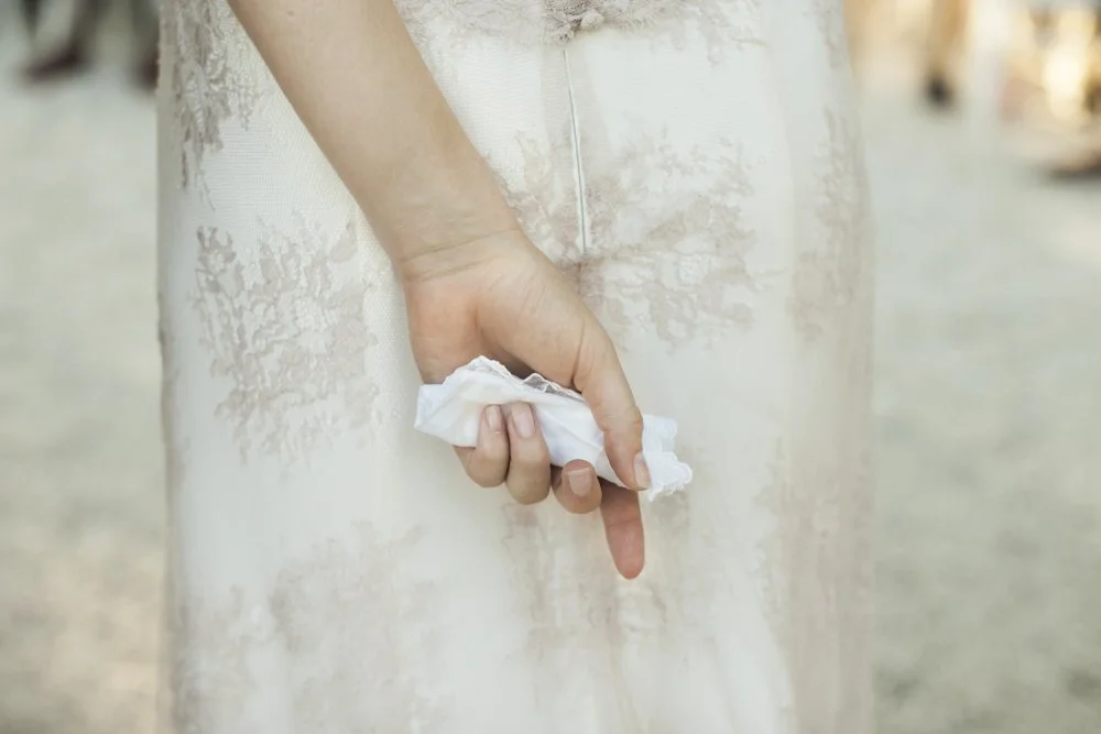 Close-up van een hand die een wit lappendje vasthoudt naast een witte kanten jurk, waarschijnlijk een trouwjurk.