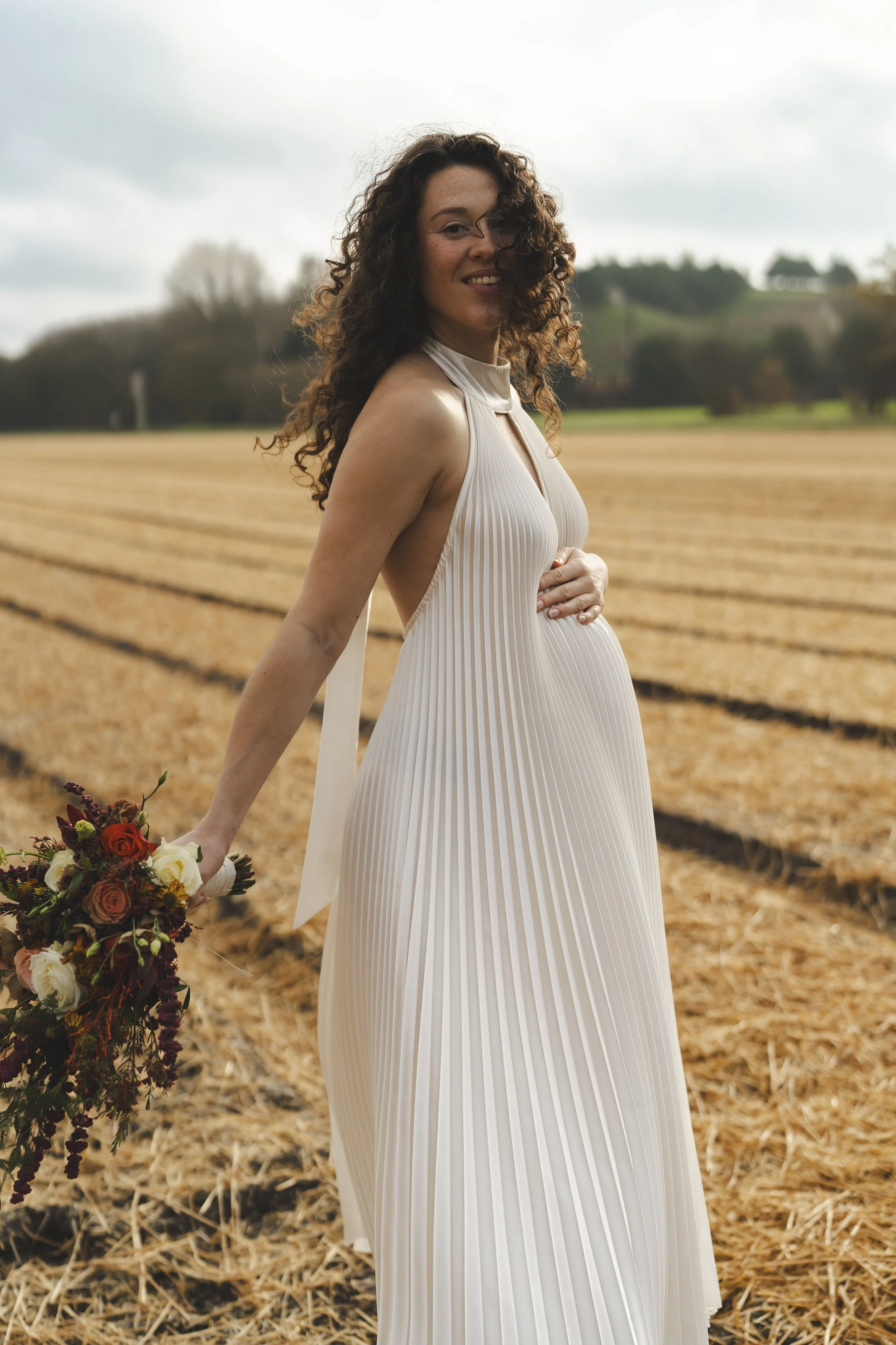 Een zwangere vrouw in een witte, plissé jurkje staat in een droog veld met een bos in de achtergrond, terwijl ze glimlacht en een bloemenboeket vasthoudt.