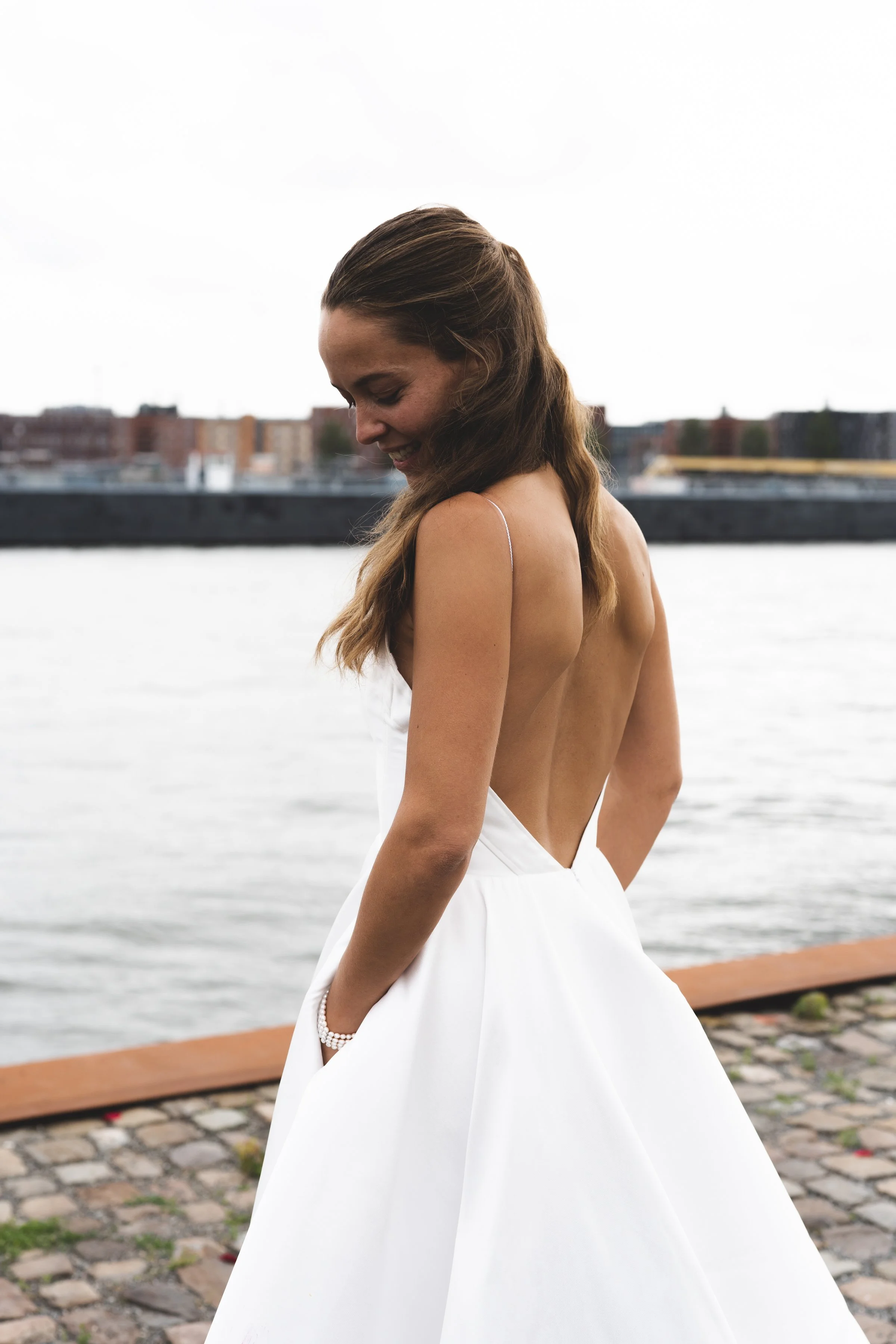 Vrouw in een witte trouwjurk met open rug, die aan een rivier staat en glimlacht.