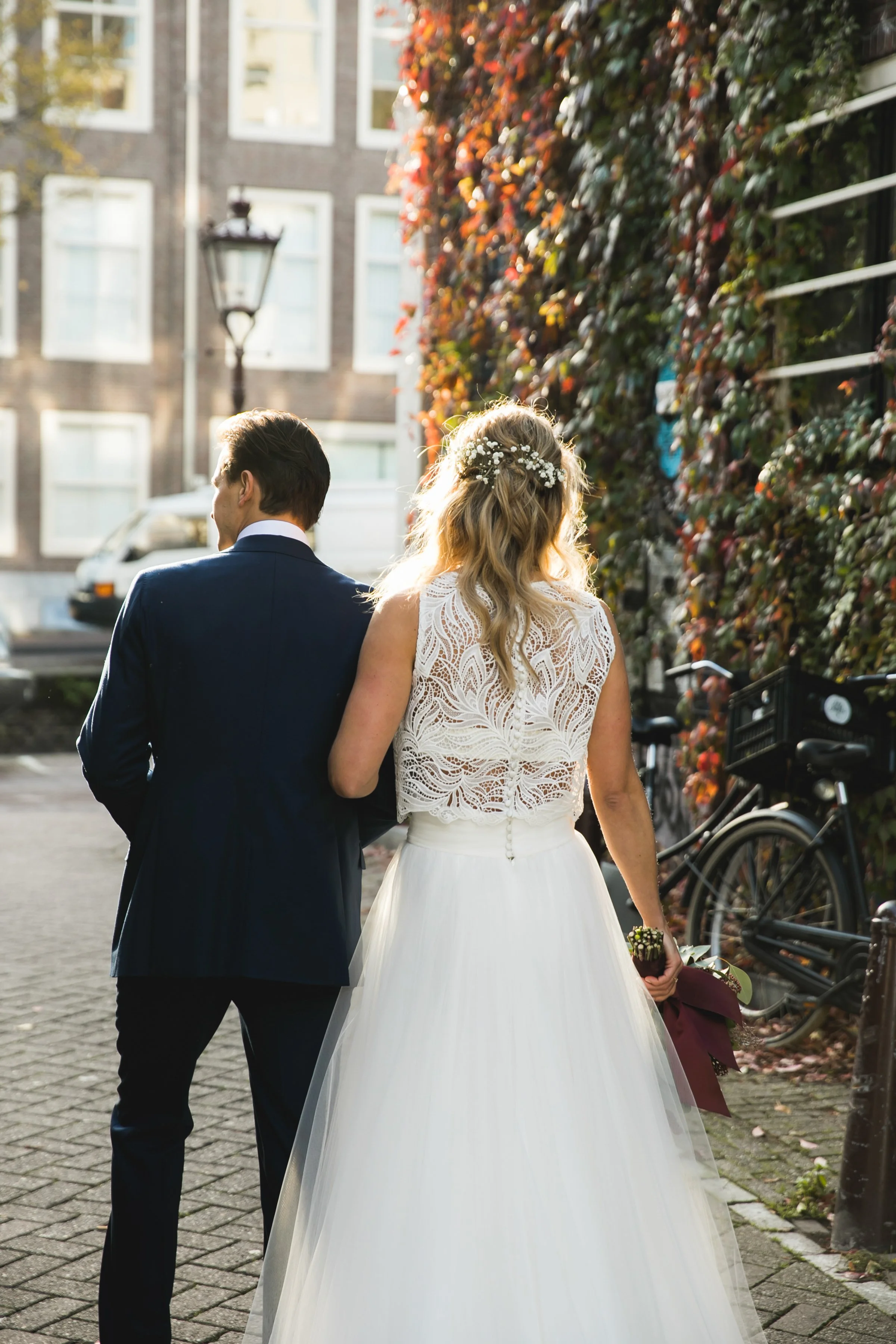 Een stel in een trouwoutfit wandelt samen op een voetpad, een man in een donkerblauw pak en een vrouw in een witte trouwjurk met kant. Ze zijn omringd door herfstkleuren, met een muur vol klimop en fietsen op de achtergrond.