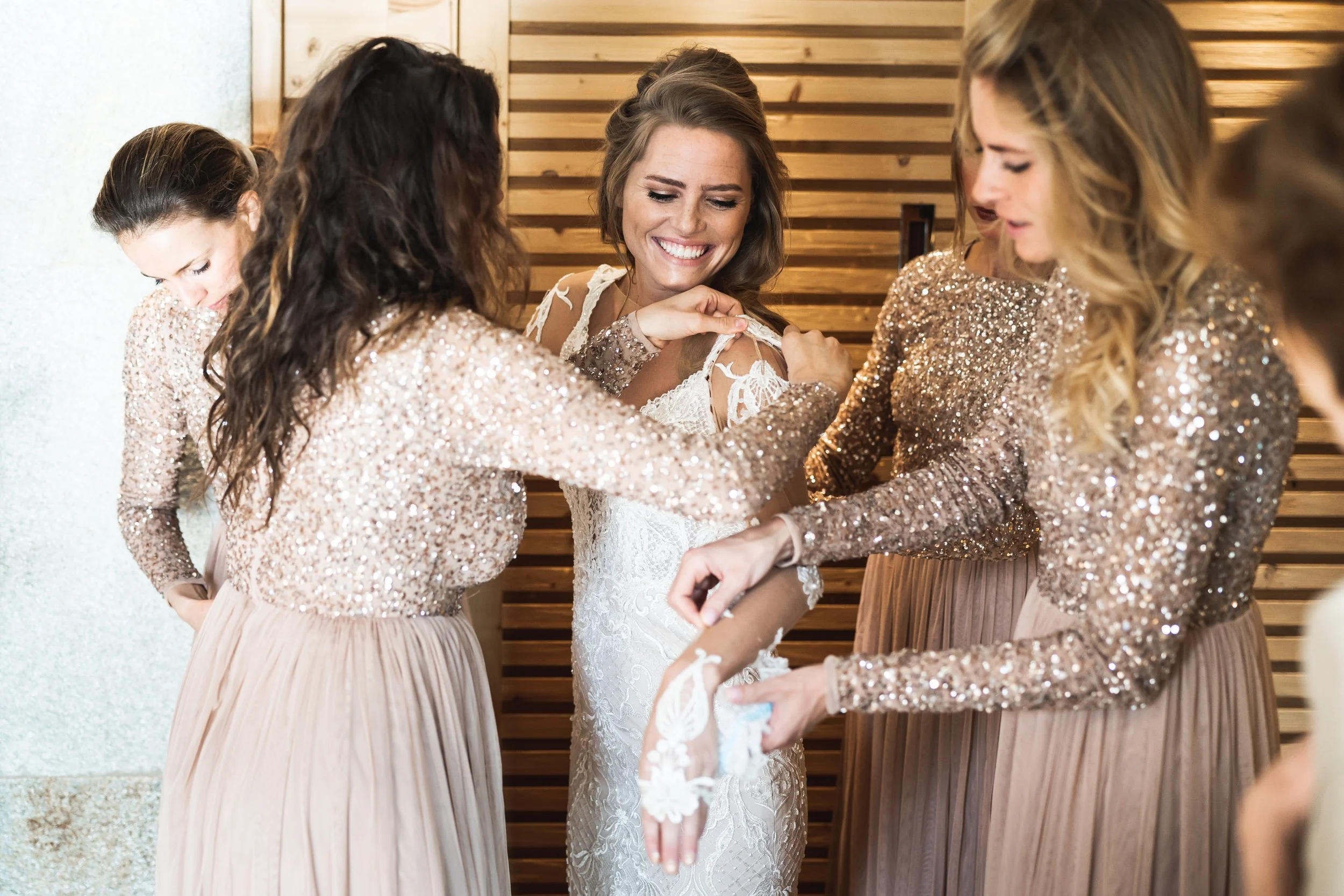 Vrouw in witte bruilofsjurk wordt geholpen door bruidsmeisjes in glitterjurk, lachend en lachend in een houten achtergrondkamer.