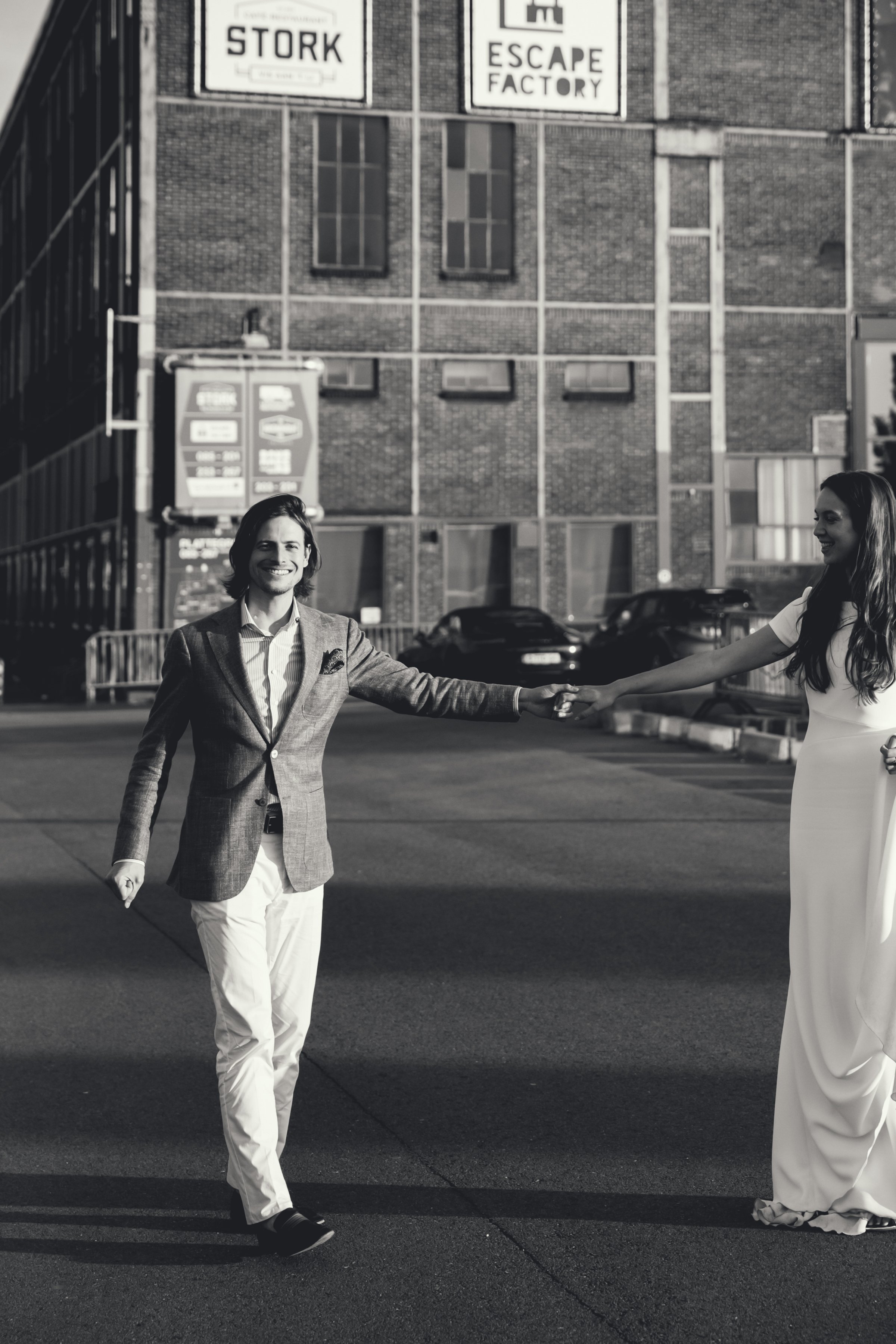 Twee mensen, een man en een vrouw, die elkaar de hand schudden op straat, met een groot gebouw op de achtergrond en parkeerplaatsen. De man draagt een pak met een colbert en de vrouw een lange witte jurk.