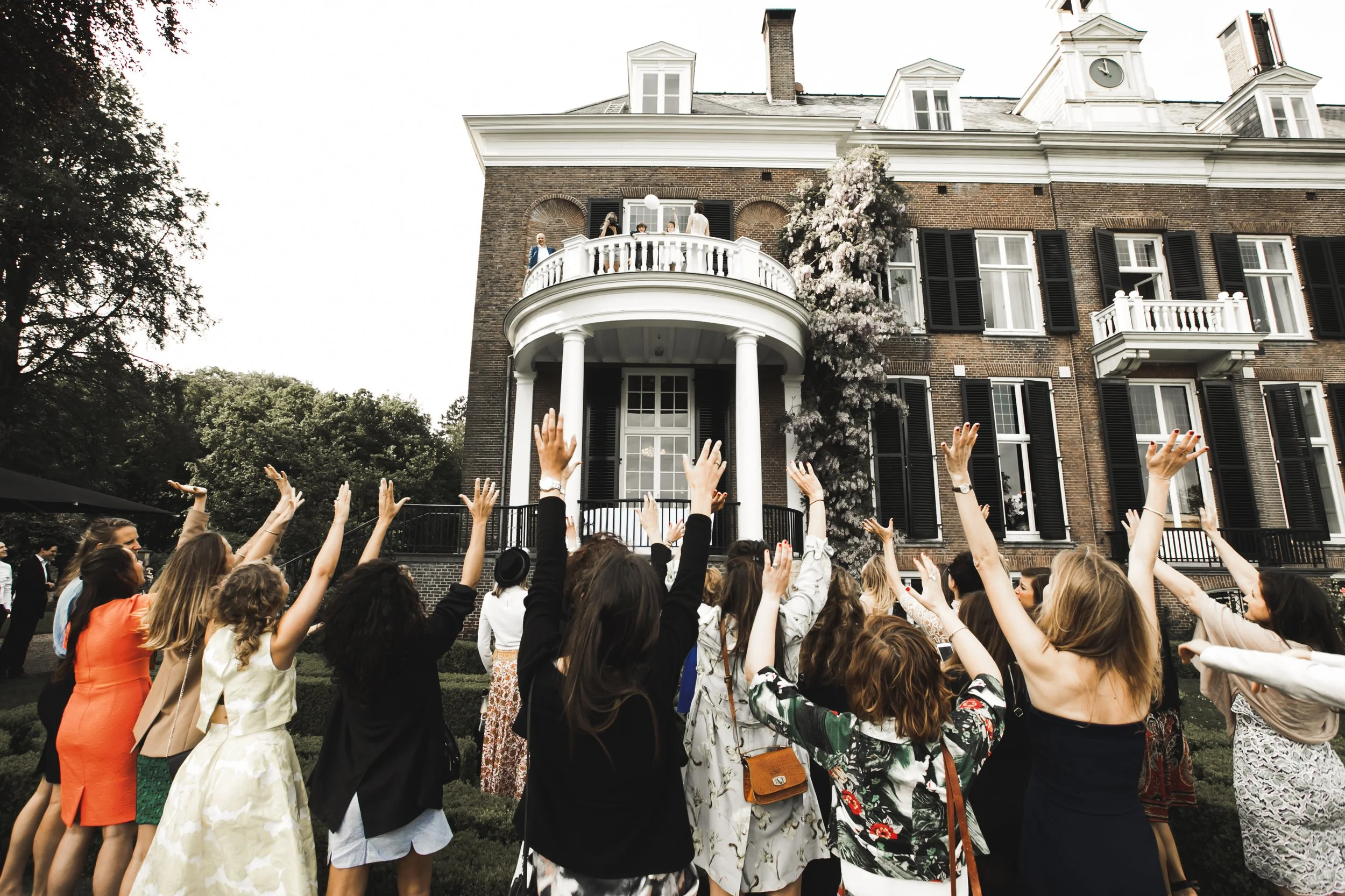 Mensen vieren een feest buiten voor een groot herenhuis, met sommigen die omhoog wijzen en anderen die op de veranda staan.