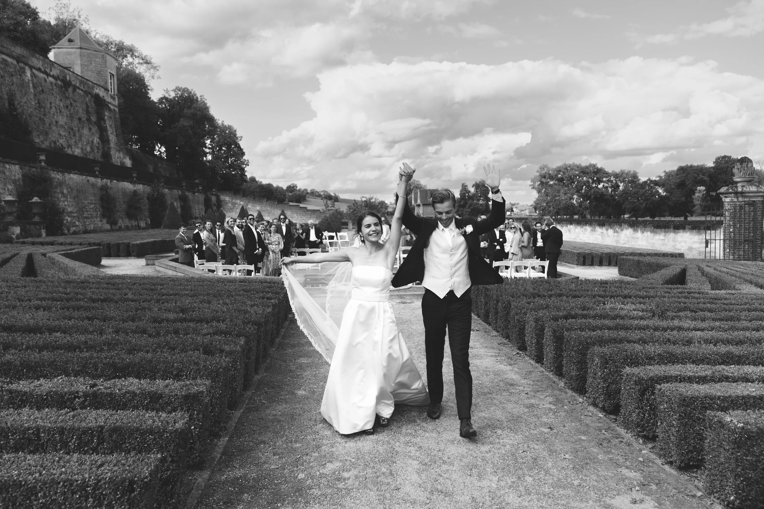 Een gelukkige bruid en bruidegom lopen hand in hand door een tuin na hun huwelijk, omringd door gasten.