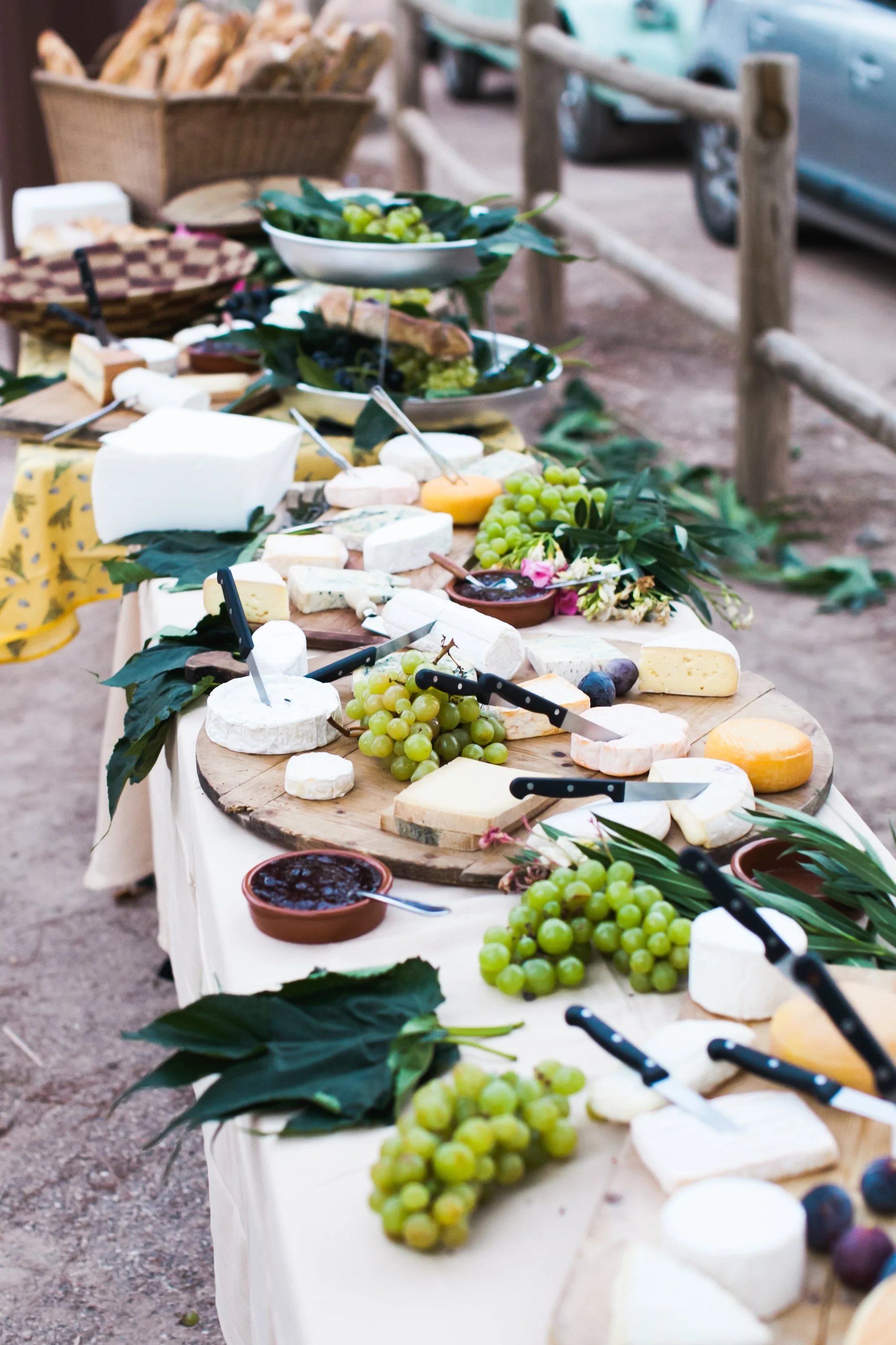 Een lang buffet met verschillende soorten kazen, druiven, jam en groenterolletjes op houten snijplanken en borden, buiten opgesteld.