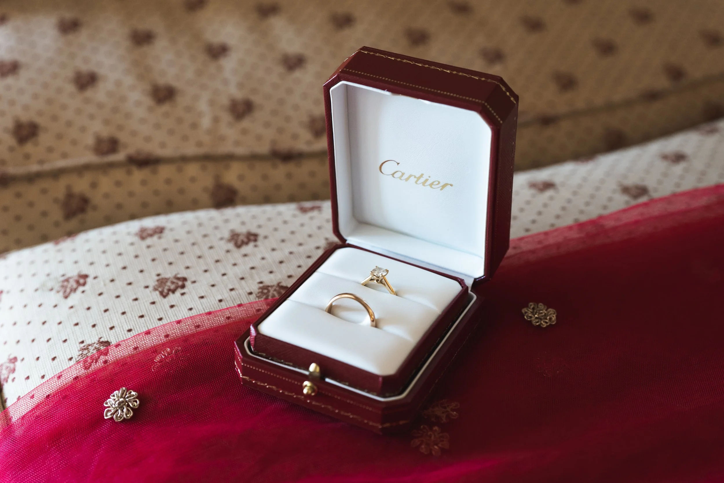 Een Cartier ring en een diamant verlovingsring in een rode sieradendoosje, geplaatst op een rood en wit stoffen oppervlak met kleine bloemen en decoratieve elementen.