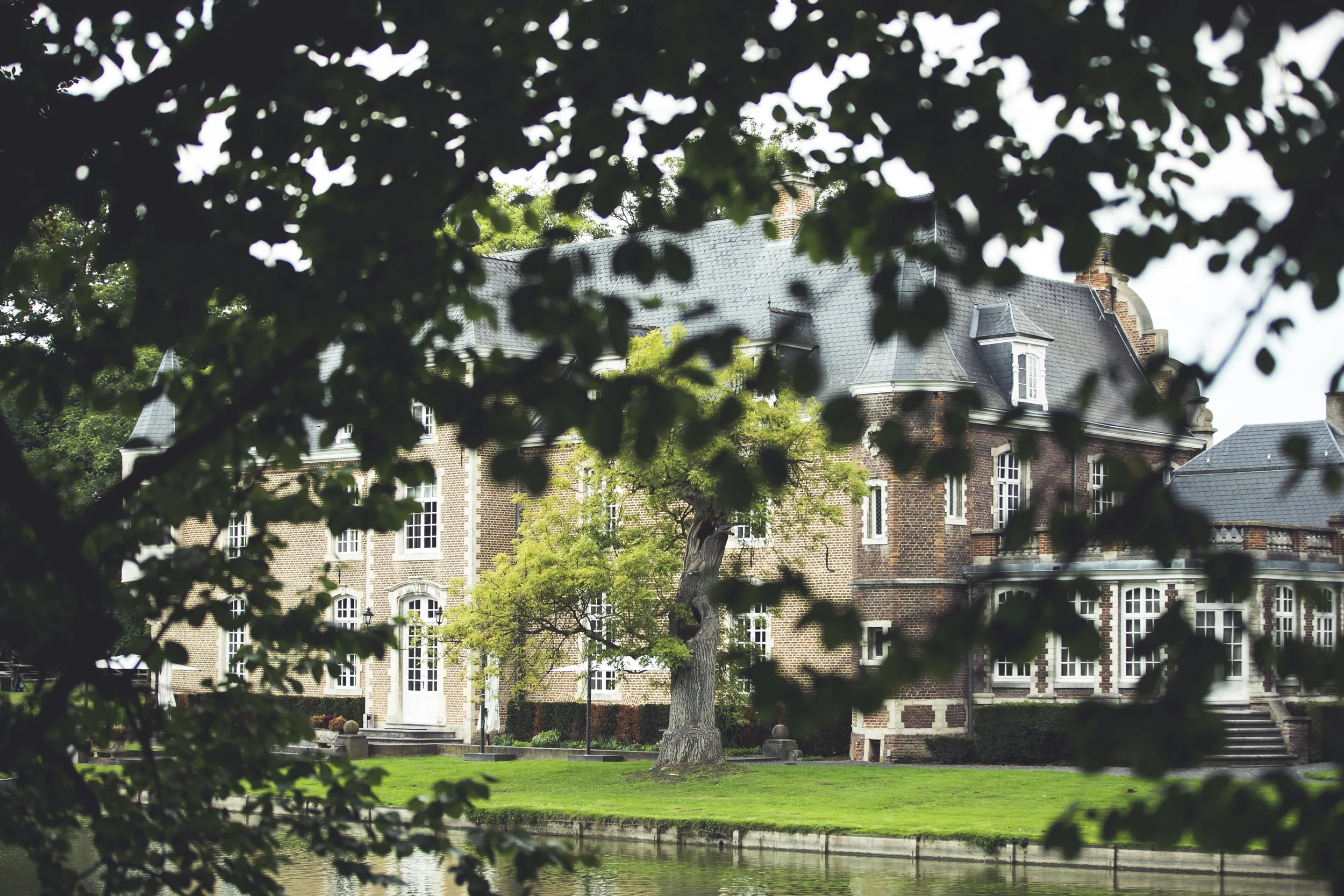 Een groot, oud, stenen herenhuis met meerdere verdiepingen en grote ramen, omgeven door groene bomen en een gazon, gezien vanaf de rand van een kleine rivier of vijver.